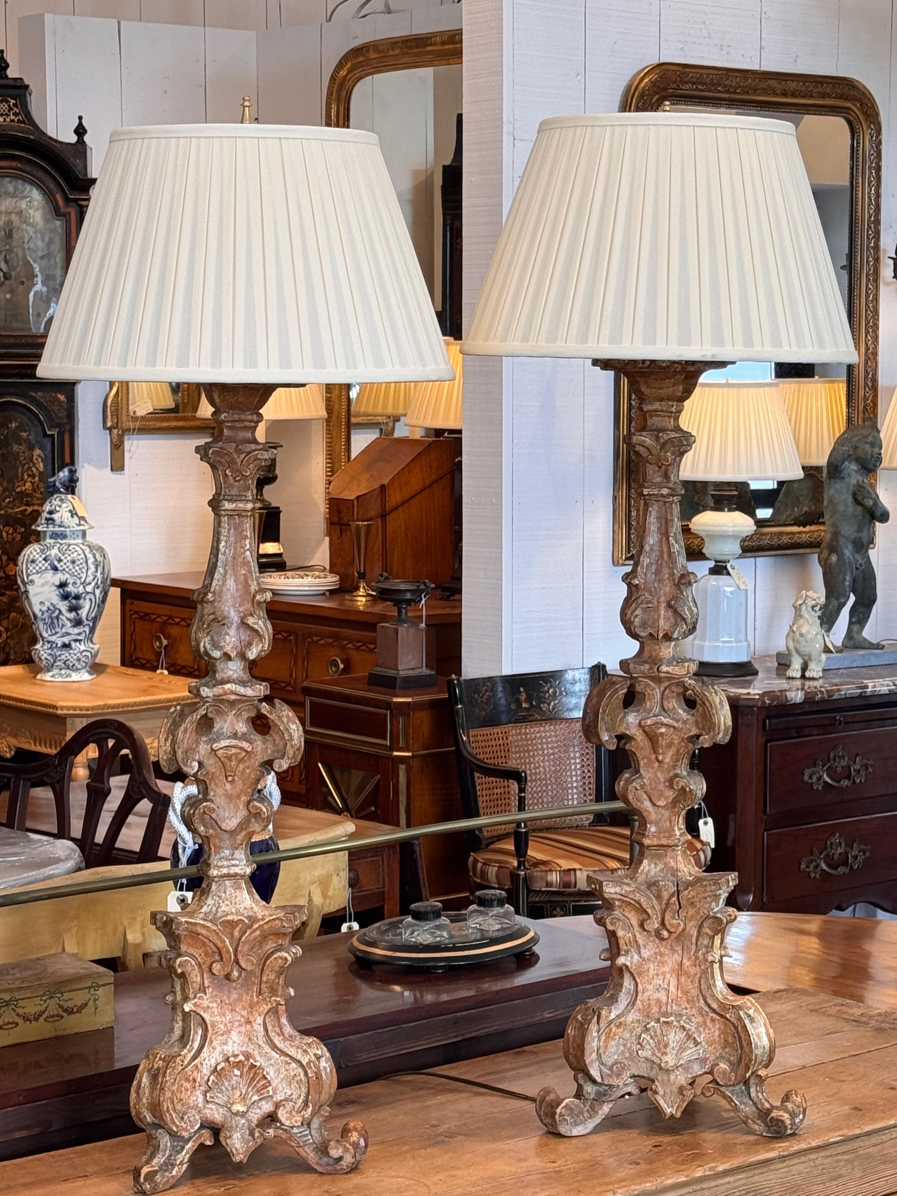 Un par de lámparas de madera dorada tallada con una elegante pátina desgastada por el tiempo. Las bases presentan intrincados detalles con hojas de acanto y motivos clásicos, inspirados en el diseño tradicional europeo. El acabado dorado envejecido