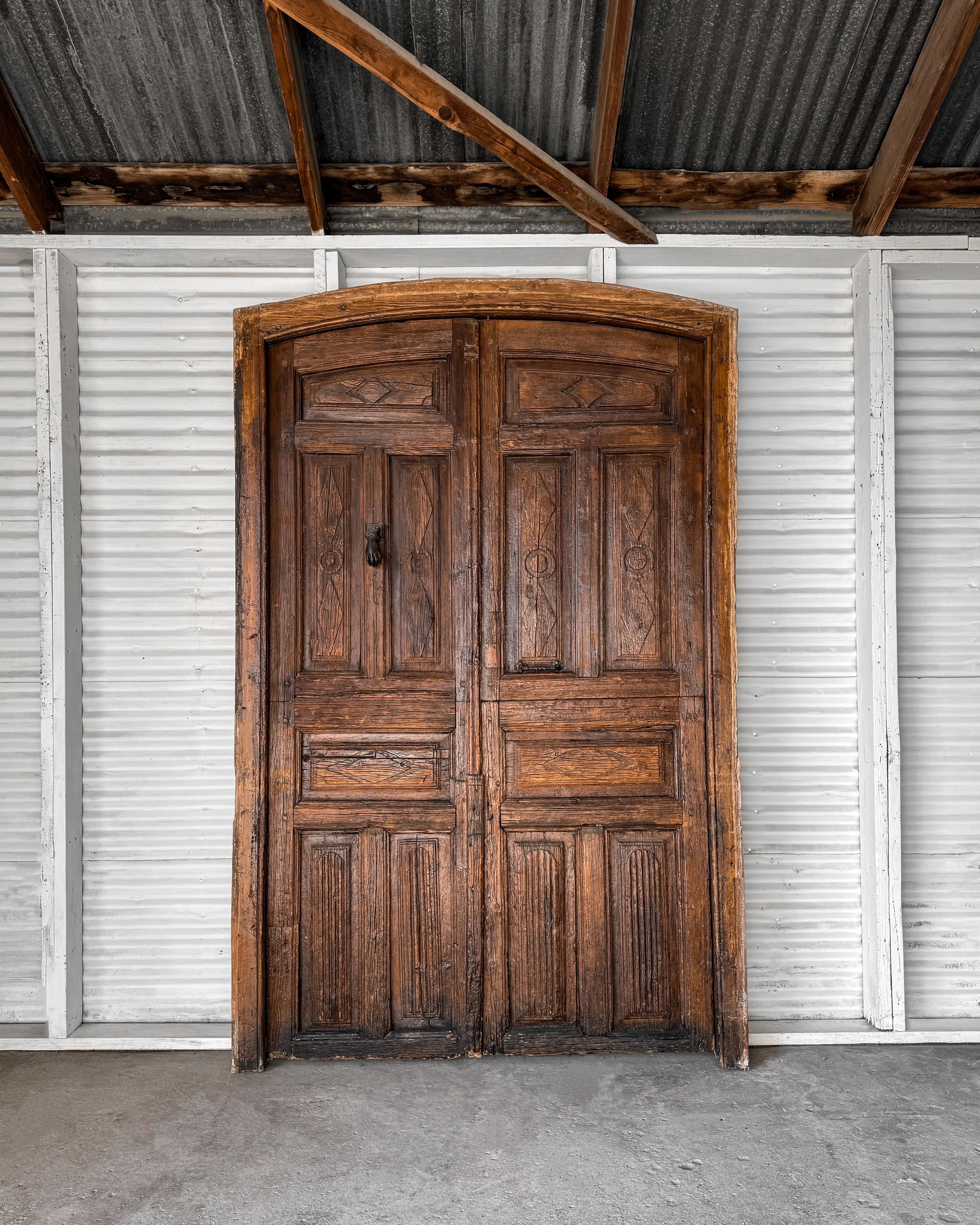 A pair of solid oak 18th-century arched paneled hacienda doors. Hand-carved details decorate the doors, which are set in the original arched frame. Both doors and frame feature a medium-dark stain that has worn to a beautiful patina. The original