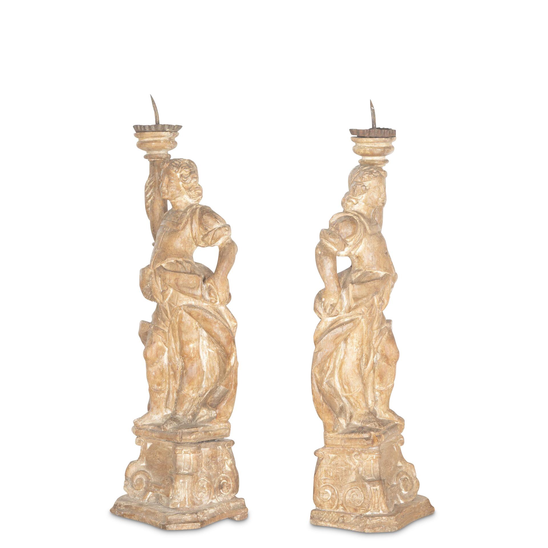 ﻿ A decorative pair of C18th Italian carved lime wood figural candelabra in classical clothing with one arm holding aloft a candle support with metal drip trays, the figures stood on scrolled plinth bases with concave mouldings. Circa 1760.

H: 72