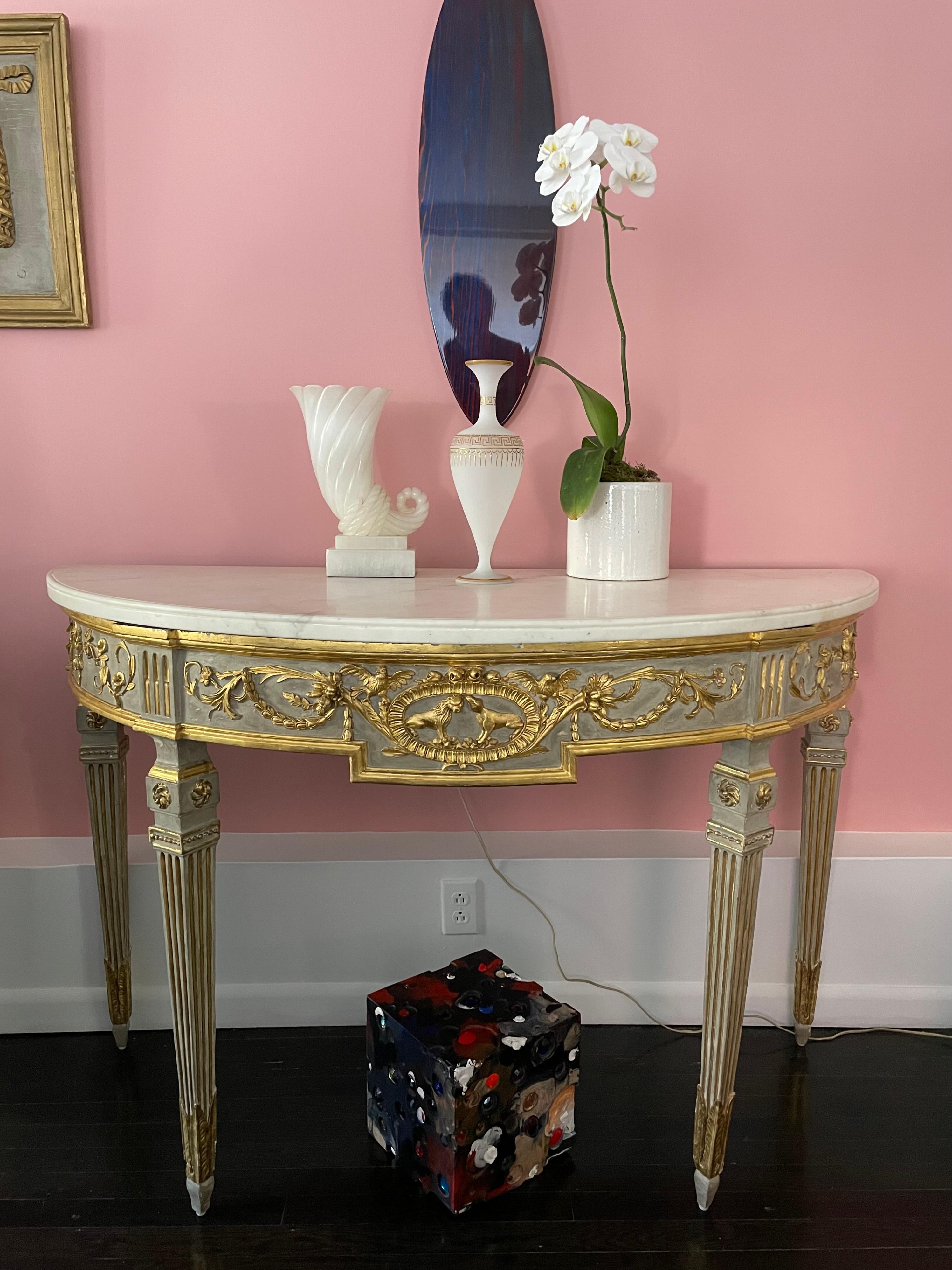 This pair of Italian Neo-Classical console tables were carved, painted battleship-grey, gilded, and topped with slabs of Carrara marble. Each has a carved cartouche — one with a pair of billy goats and the other with a lion and lioness — flanked by