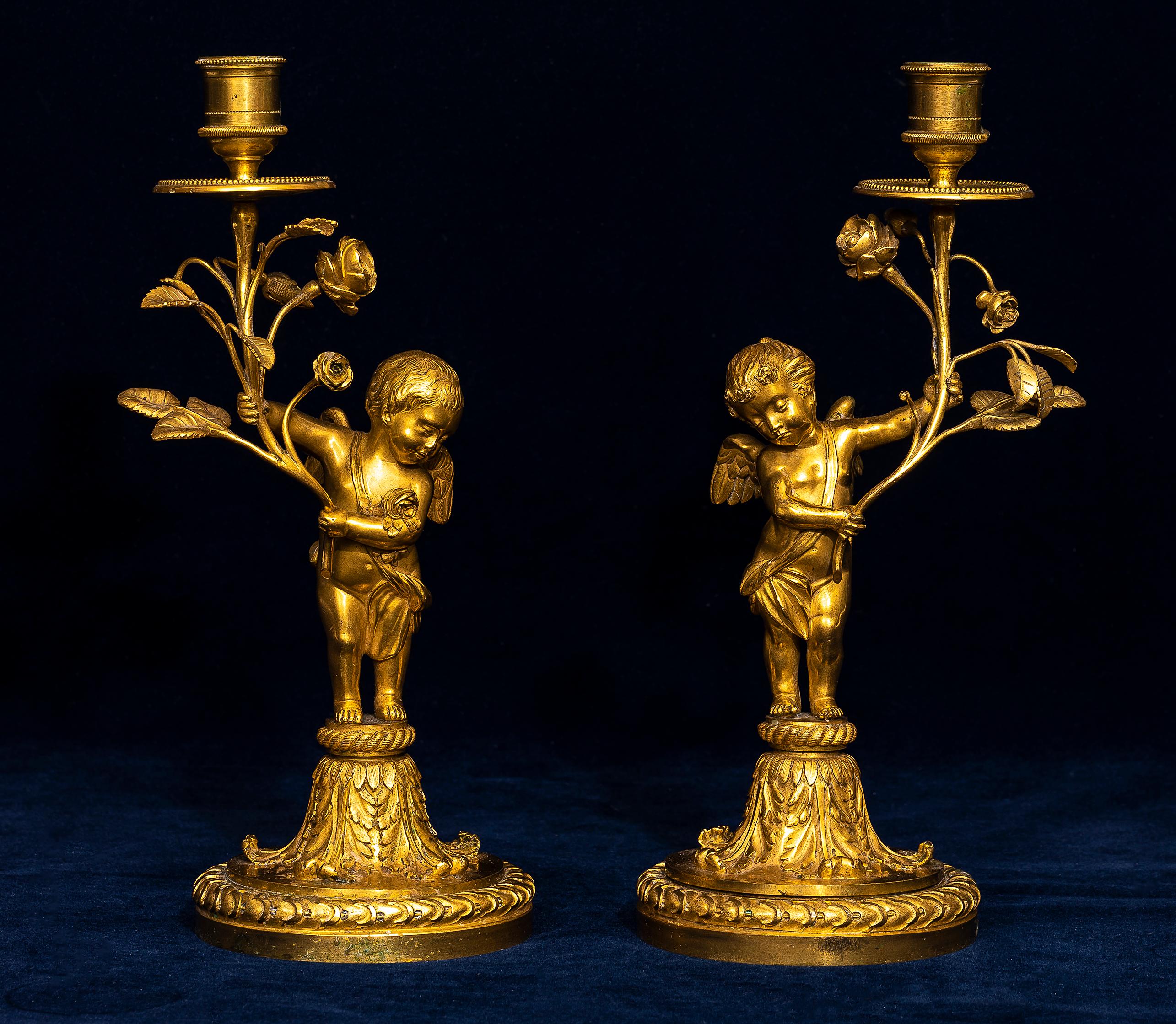 A pair of 18th century Louis XVI ormolu candlesticks, each modeled as a standing cupid holding a flowering rose branch supporting the single candleholder. The figures are cast in full round, with feathered wings, defined facial features, and softly