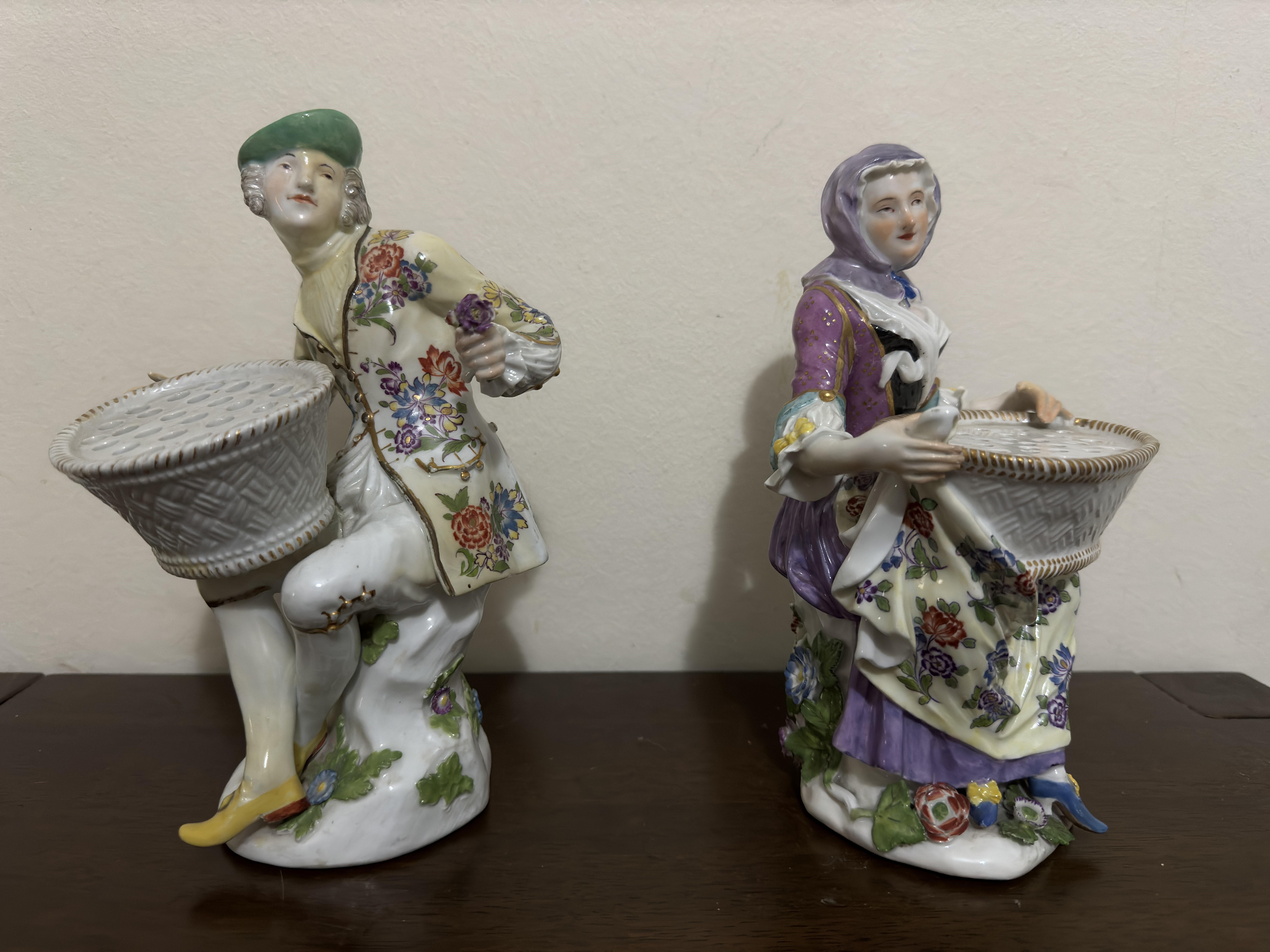 A pair of Meissen bouquetiere figures Circa 1765, both with 'Christie's East' dealer stamps.
Modelled by J.J. Kändler holding oval two-handled baskets, the tops pierced for flowers, he in green cap and white breeches, she in purple scarf, pink