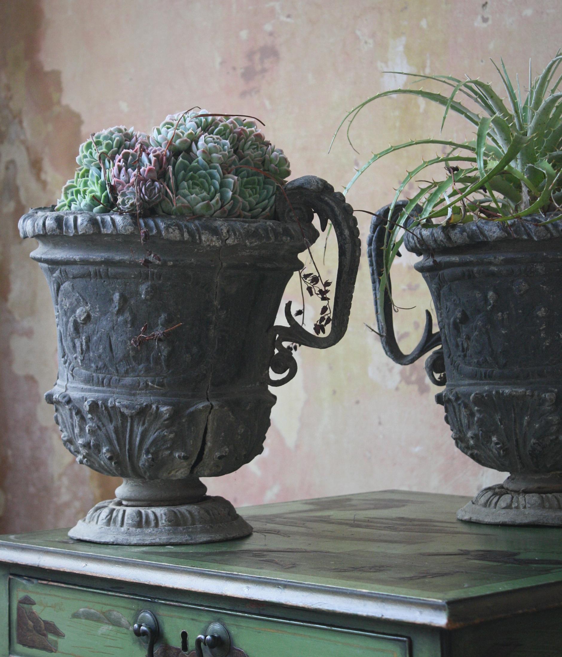 A highly decorative matching pair of English lead urns in the 18th century style.

Typical scrolled handles and gadrooned bases, classical figures adore the central body of each urn.

Some sagging and minor splitting as one would expect due to