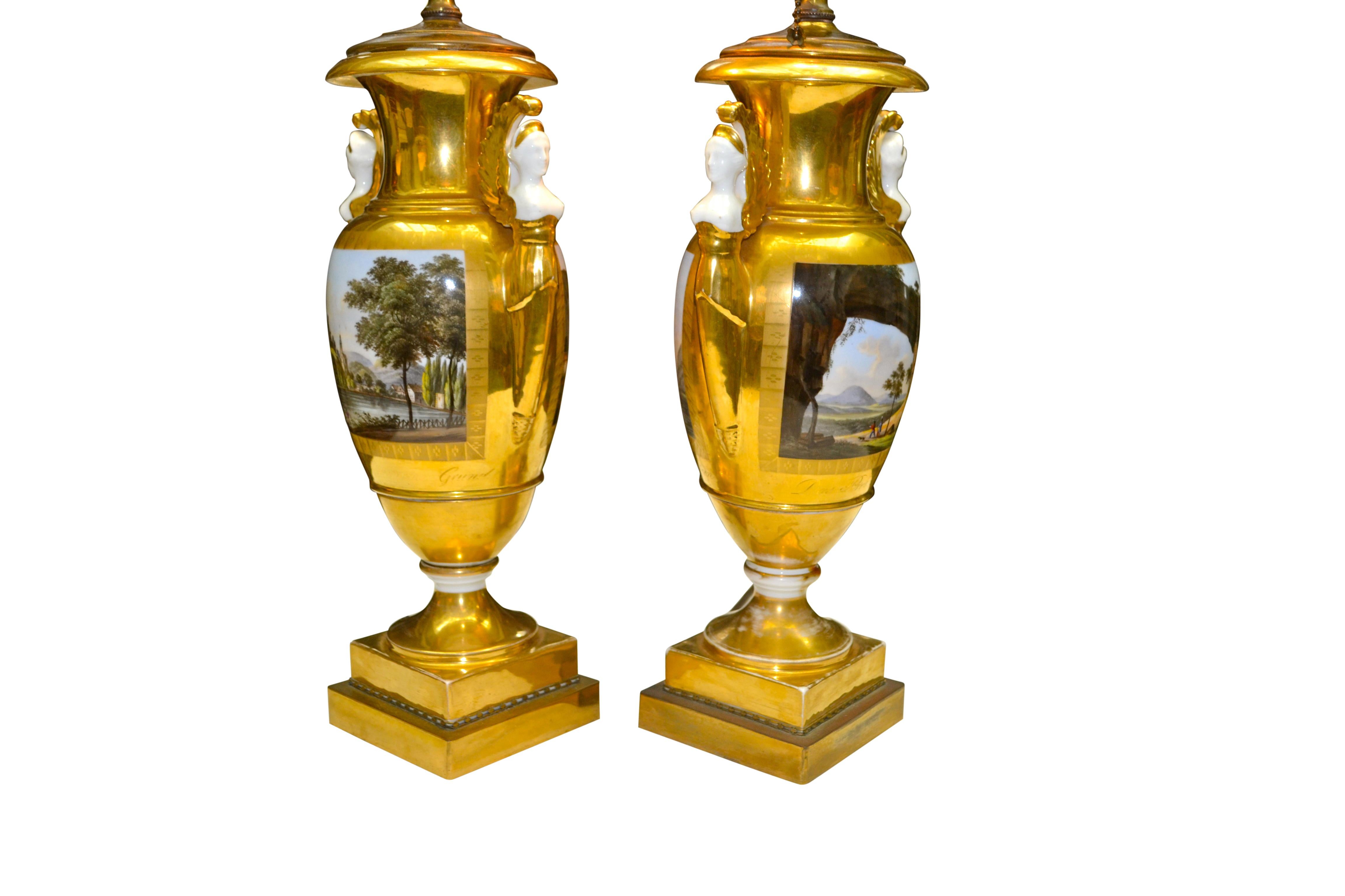 The twin handled urn shaped porcelain body is entirely gilded with matte and burnished gold. Each partially gilded upper ‘handle’ is in the shape of a winged classical female torso. The urns are painted on each side with familiar German landscapes