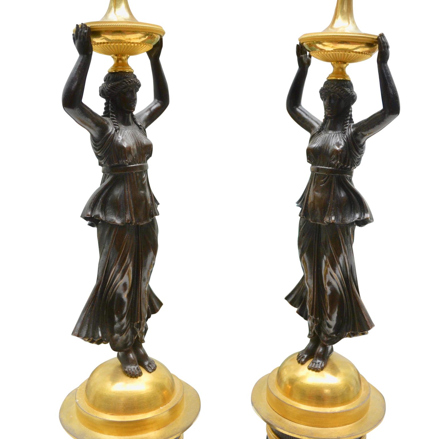 A pair of Russian Empire figural candelabra featuring a patinated bronze caryatid holding a gilt bronze urn mounted with a torch atop her head, with two horns terminating in flamed candleholders, the whole raised on a gilt and patinated bronze domed