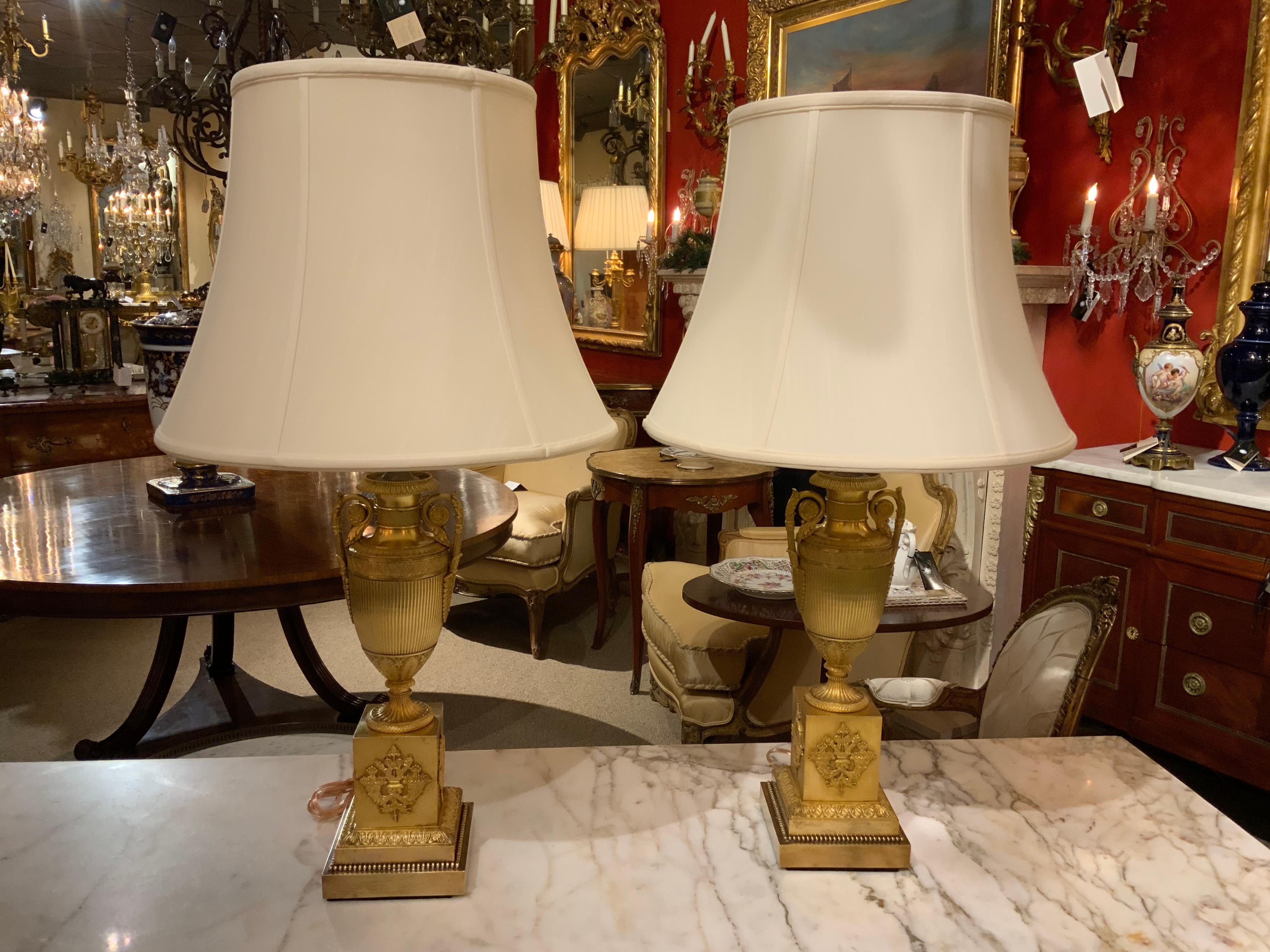 Exceptional gilding on this pair of urns that have been converted into lamps.
The urns having a curved handle on either side of the baluster form urn.
The urn has a reeded design and is set on a square base with a design
Of a torch and quiver