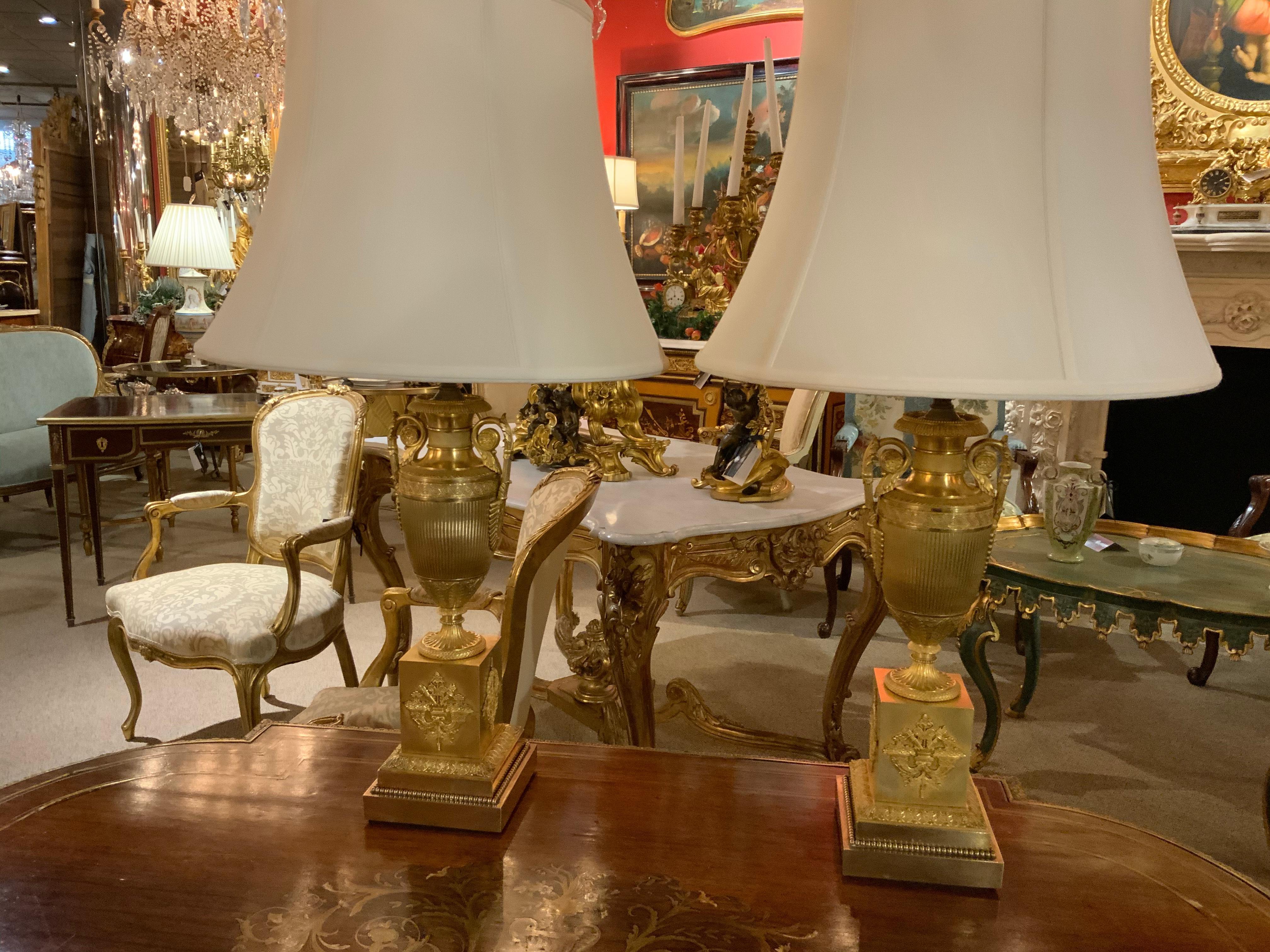 Exceptional gilding on this pair of urns that have been converted into lamps.
The urns having a curved handle on either side of the baluster form urn.
The urn has a reeded design and is set on a square base with a design
Of a torch and quiver