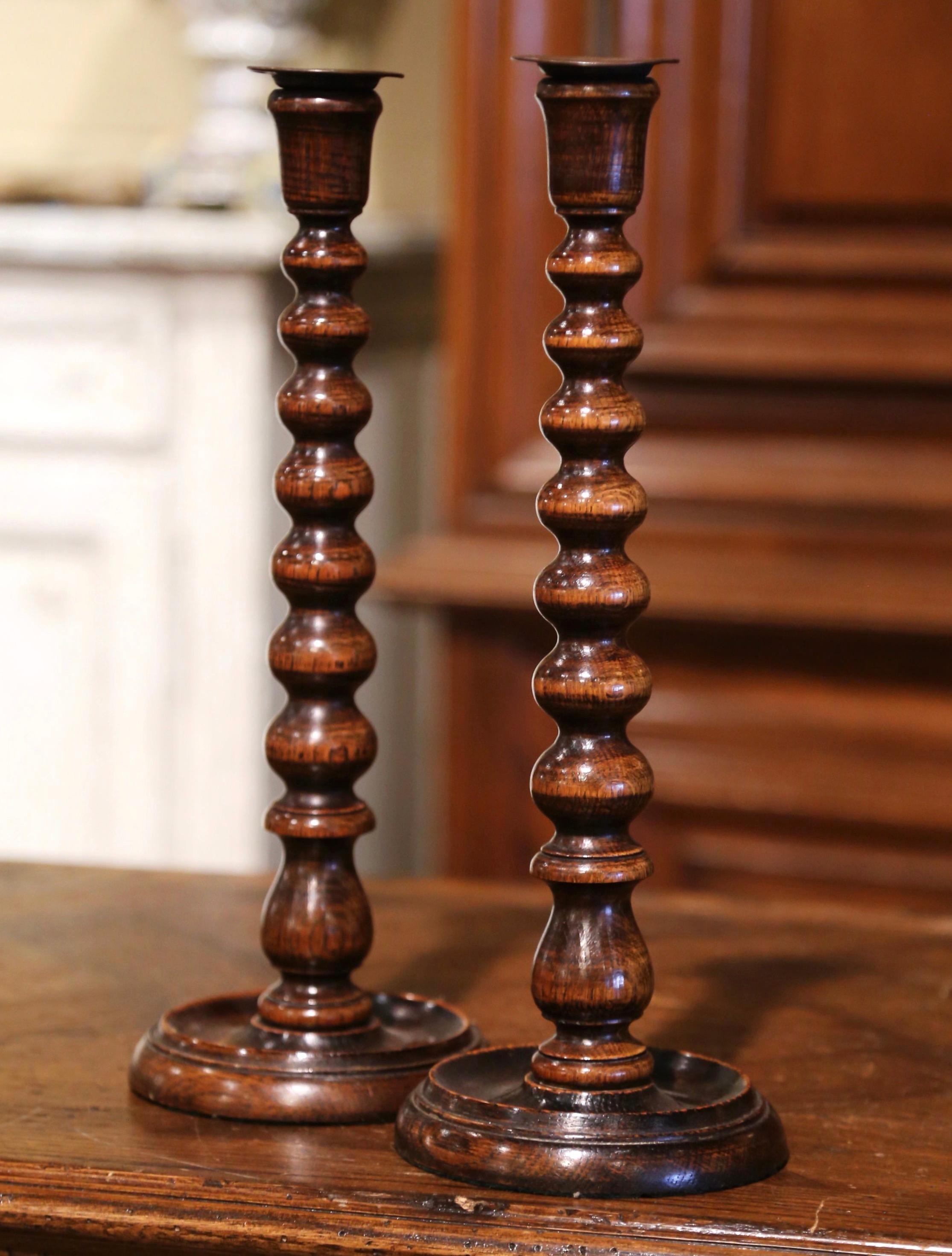 These antique candlesticks were crafted in England circa 1920. Standing on a round base, each candle holder features a open carved bobbin turned stem decorated with a brass bobeche at the top. Both tall candlesticks are in excellent condition with a