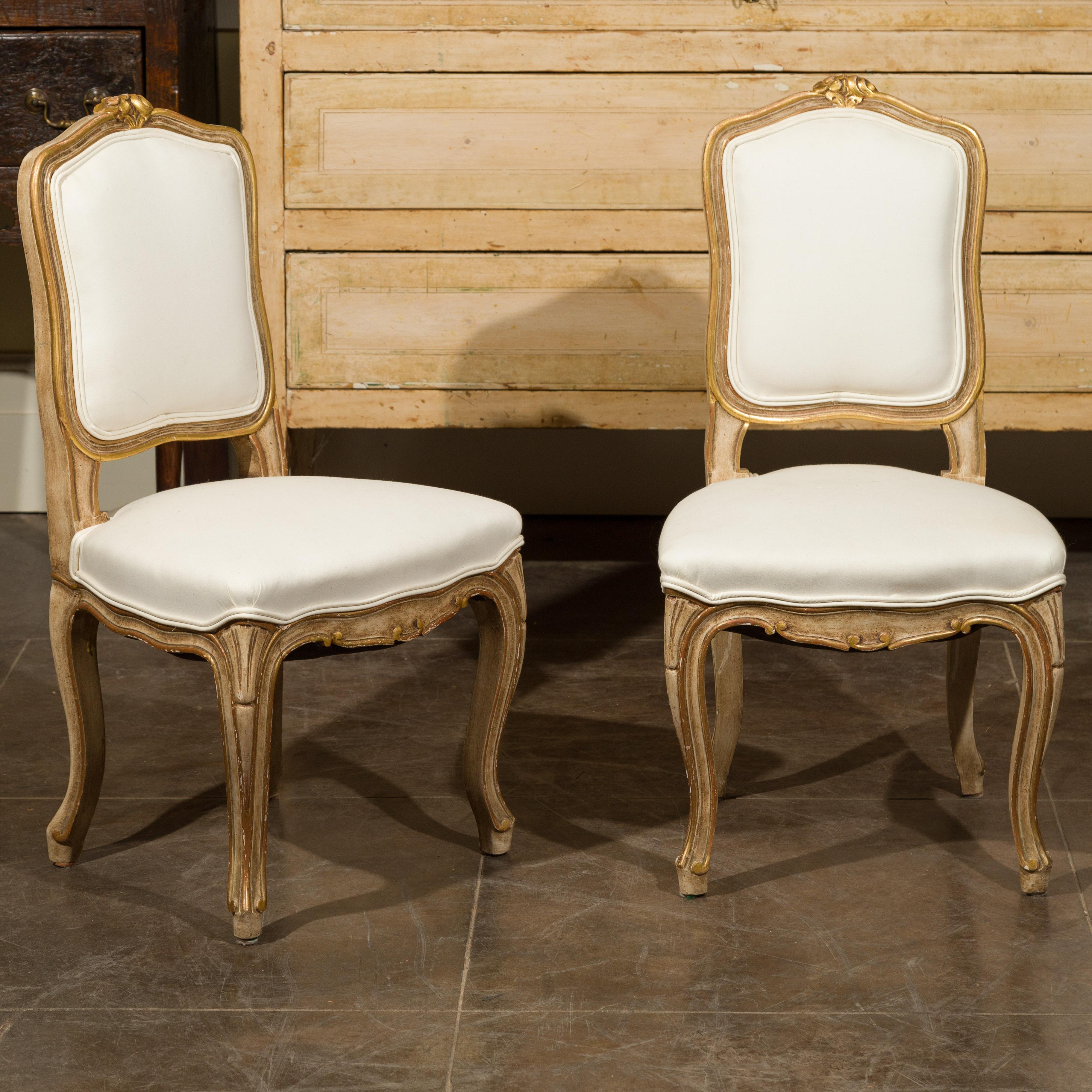 A pair of French Louis XV style painted and gilt wooden child's side chairs from the early 20th century, with carved flowers and new upholstery. Created in France during the first quarter of the 20th century, each of this pair of child's chairs