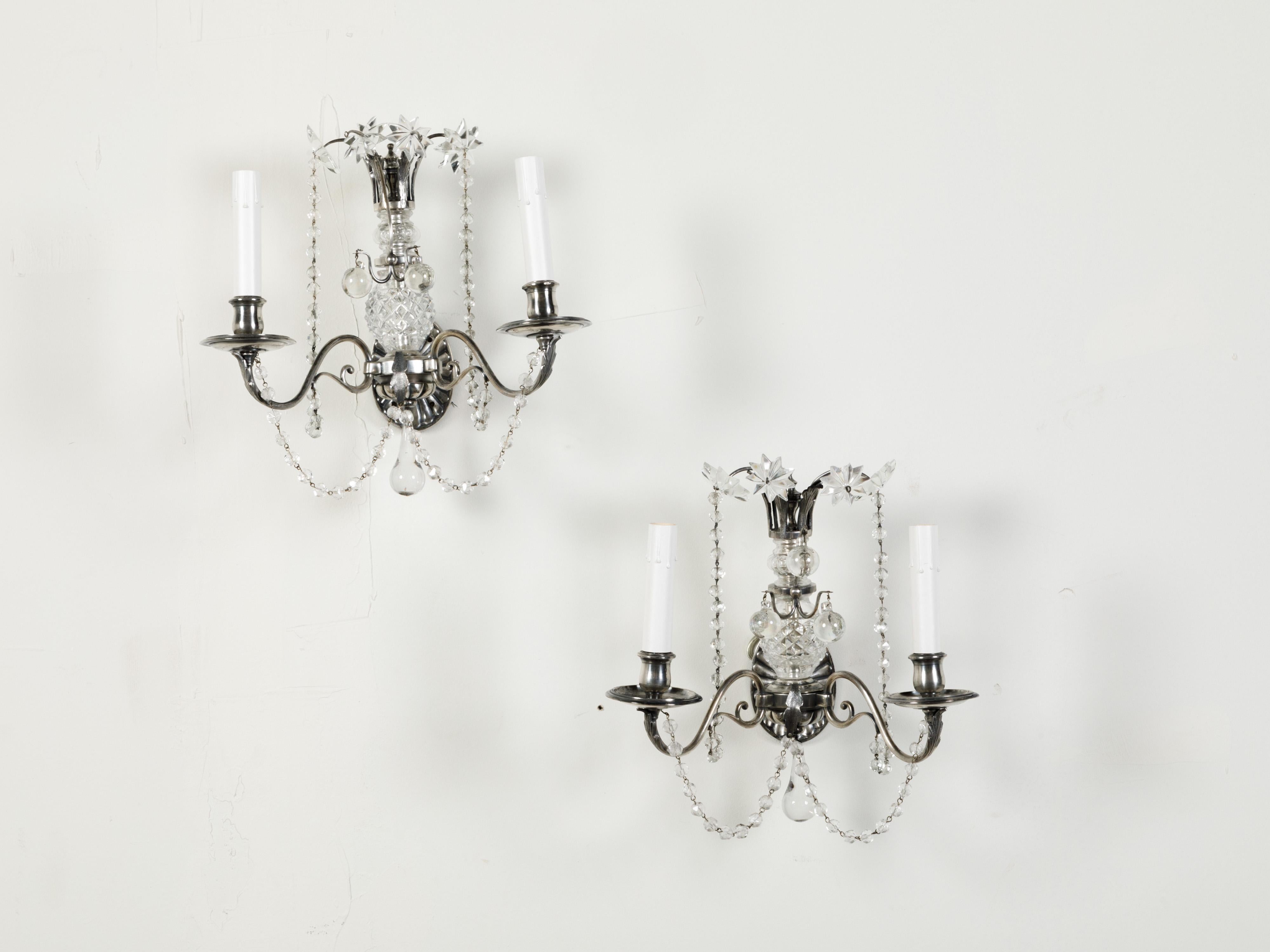 A pair of metal and crystal wall sconces from the early 20th century, with two arms, rosettes and beads. Created during the first half of the 20t century, each of this pair of crystal sconces features a radiating backplate supporting two elegant