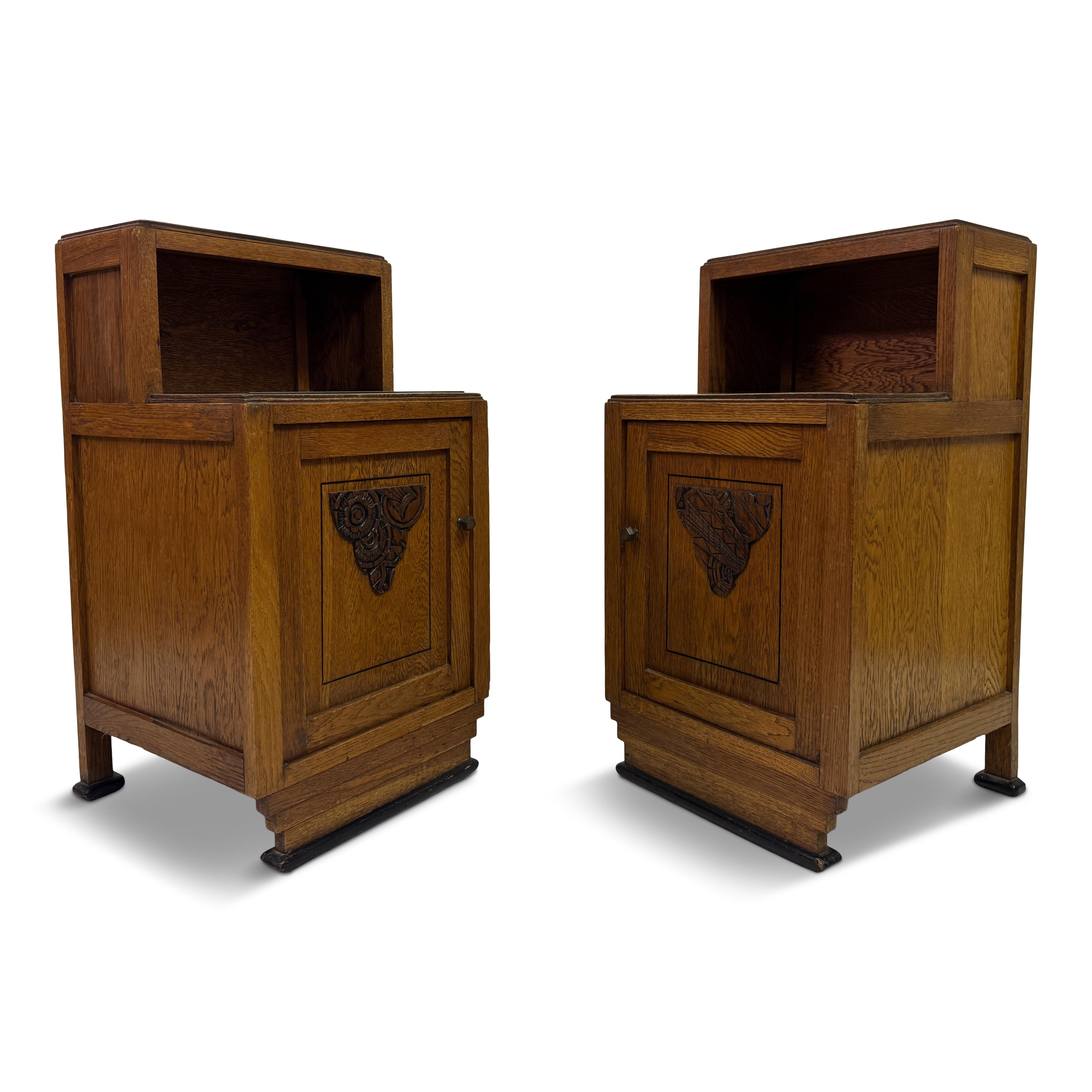 A handsome pair of bedside cabinets in oak, full of quiet character and robust charm. Each piece features a deep, warm grain and a square, architectural form softened by a touch of ornament — the cupboard doors are inset with hand-carved panels in a