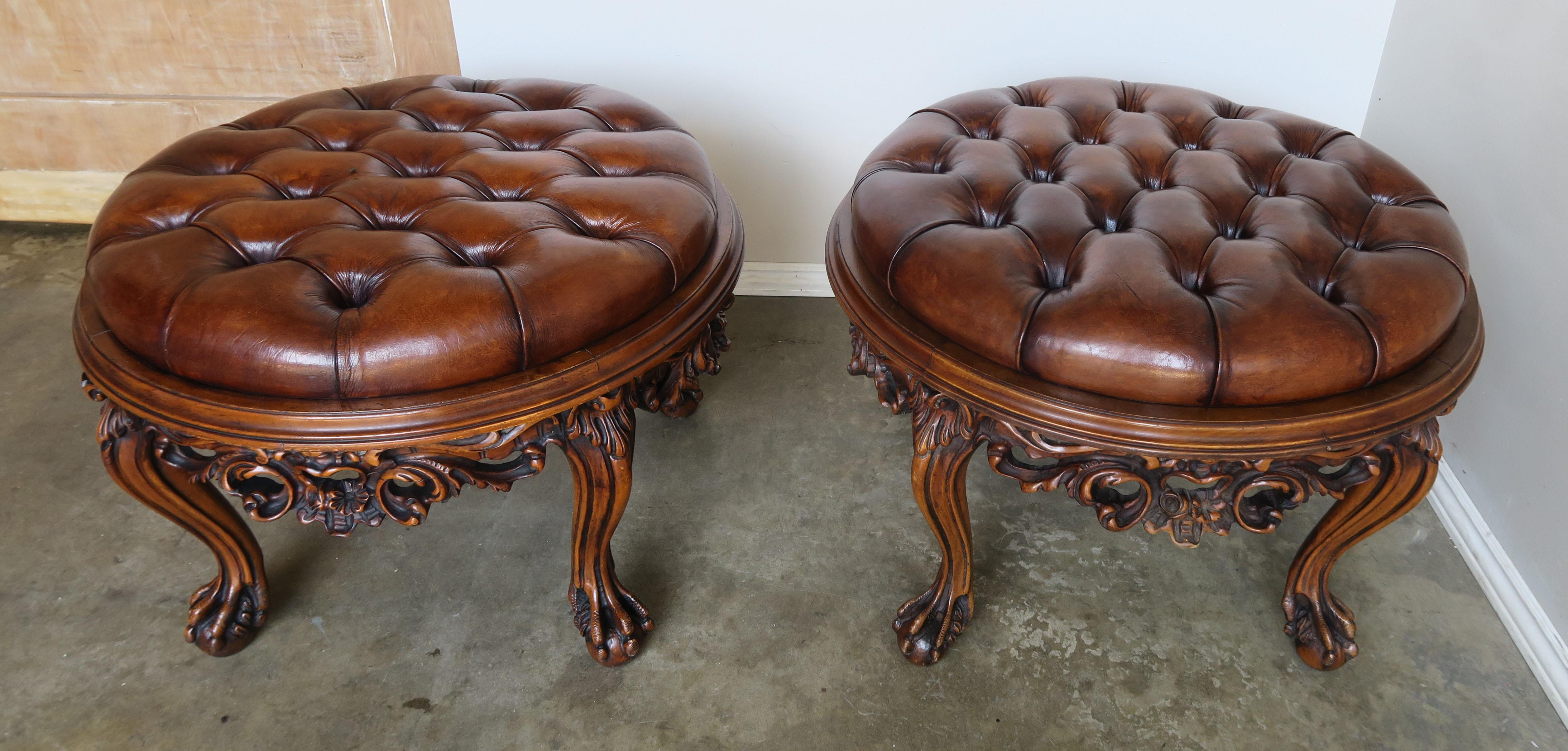 A matched pair of 1930s ornately carved French Louis XV style walnut ottomans standing on four cabriole legs that end in ball and claw feet. The round shaped pieces are upholstered in tufted leather with a beautiful distressed finish.
