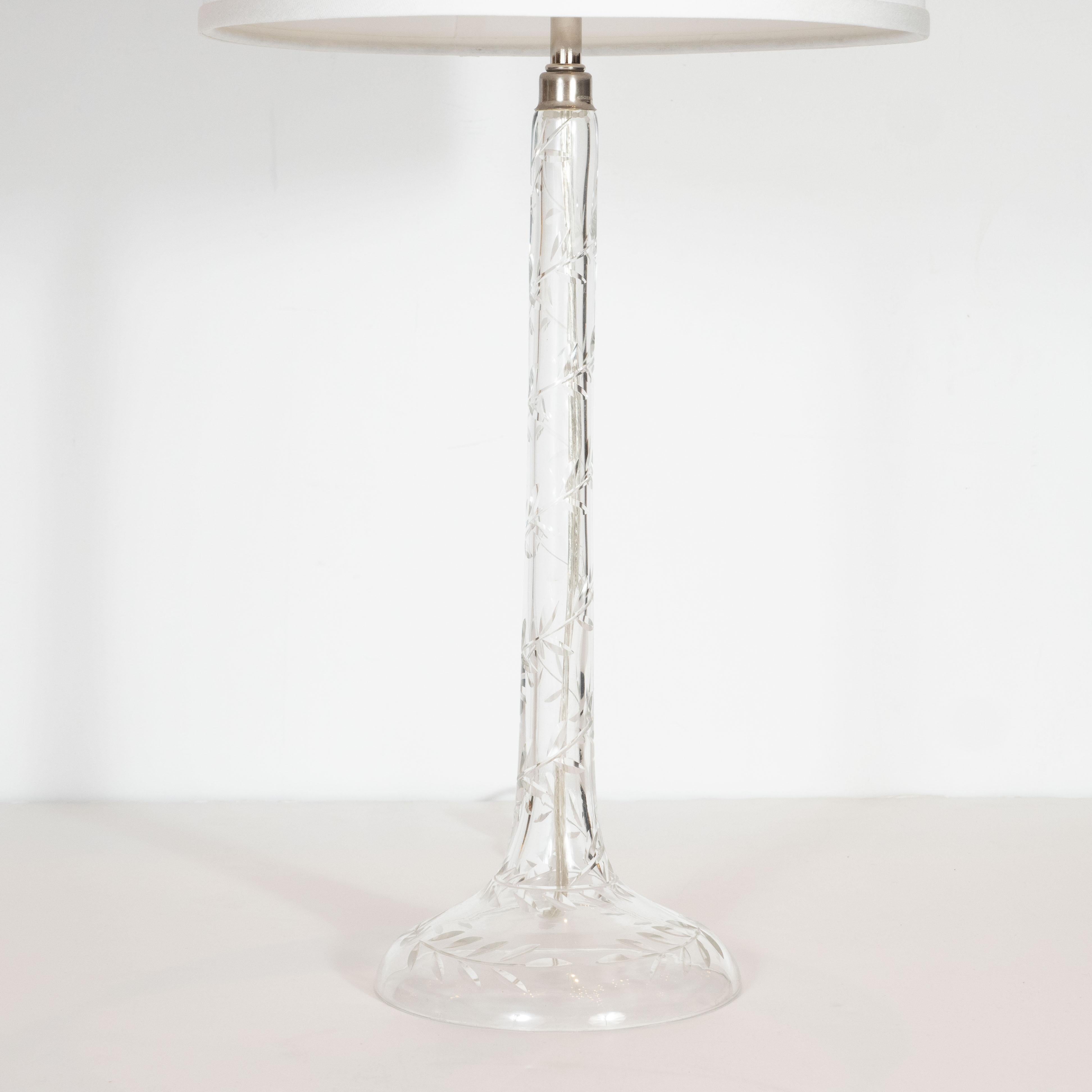 This refined and glamorous pair of Hollywood Regency table lamps were realized in the United States, circa 1940. They feature translucent crystal bodies with etched foliate designs, suggesting stylized laurel bows; an undulating brass finial; and a