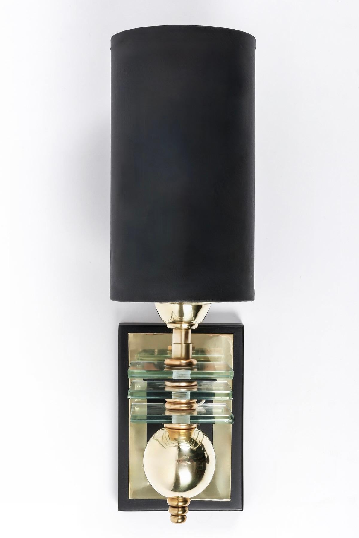 The wall base is composed of a black rectangle embellished with a gilded brass rectangle on which rests the light arm decorated with three glass plates and highlighted with brass balls on the lower part.
On the upper part, the sconce is dressed with