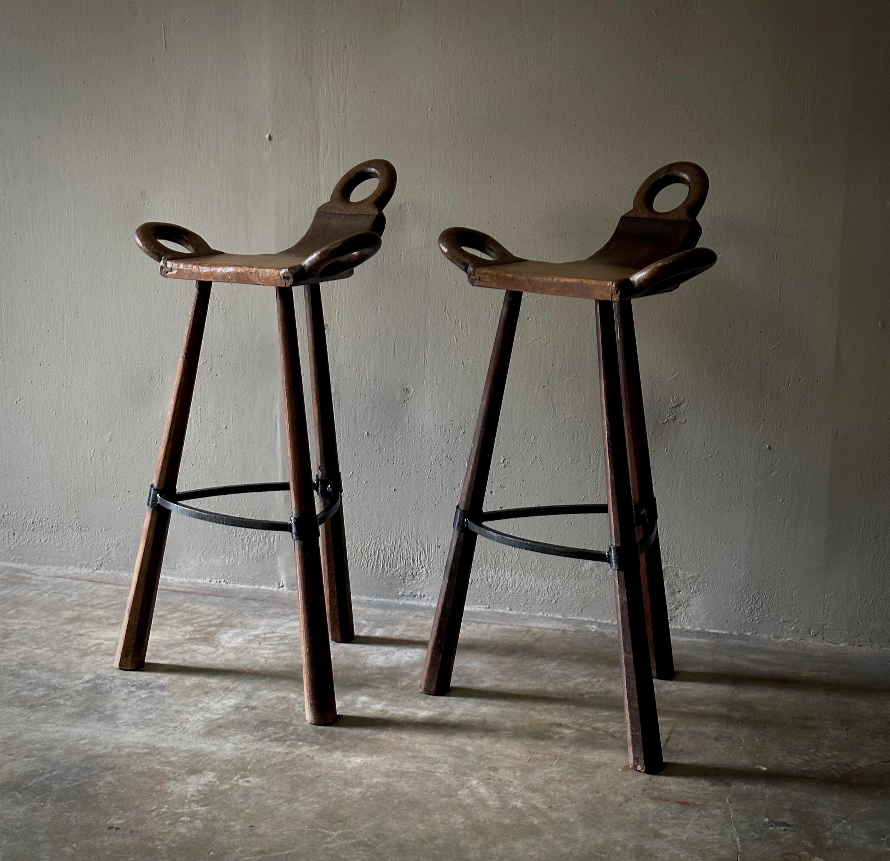 Pair of Spanish 1940s wooden workshop stools with adjoined three-legged bases, metal footrests and interesting carved ring hook accenting. Elegant and industrial with a unique silhouette. 

Spain, circa 1940.

Dimensions: 22W x 17D x 35H (seat