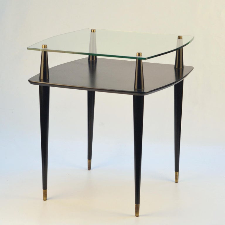 Pair of double level coffee tables with tops of tempered glass and painted wooden shelve below. The tops are connected by four black lacquered wooden tapered legs. They are highlighted by brass fittings connecting the legs and feet. This table