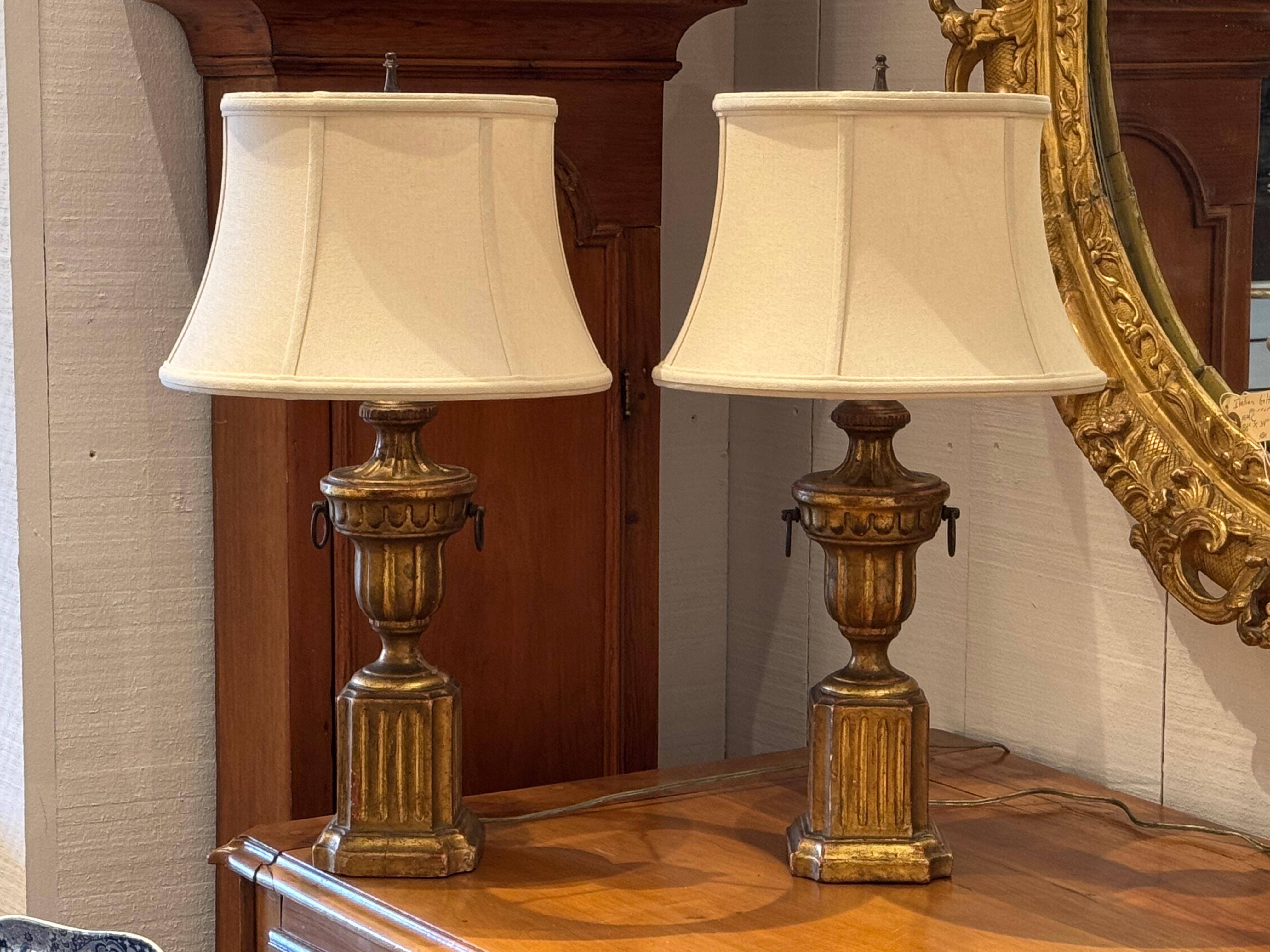 Clásica pareja de lámparas de sobremesa de estilo Borghese con base en forma de urna tallada y dorada. Las columnas estriadas y los zócalos de bloque evocan el diseño neoclásico, con un dorado desgastado que añade calidez y antigüedad. Las sutiles