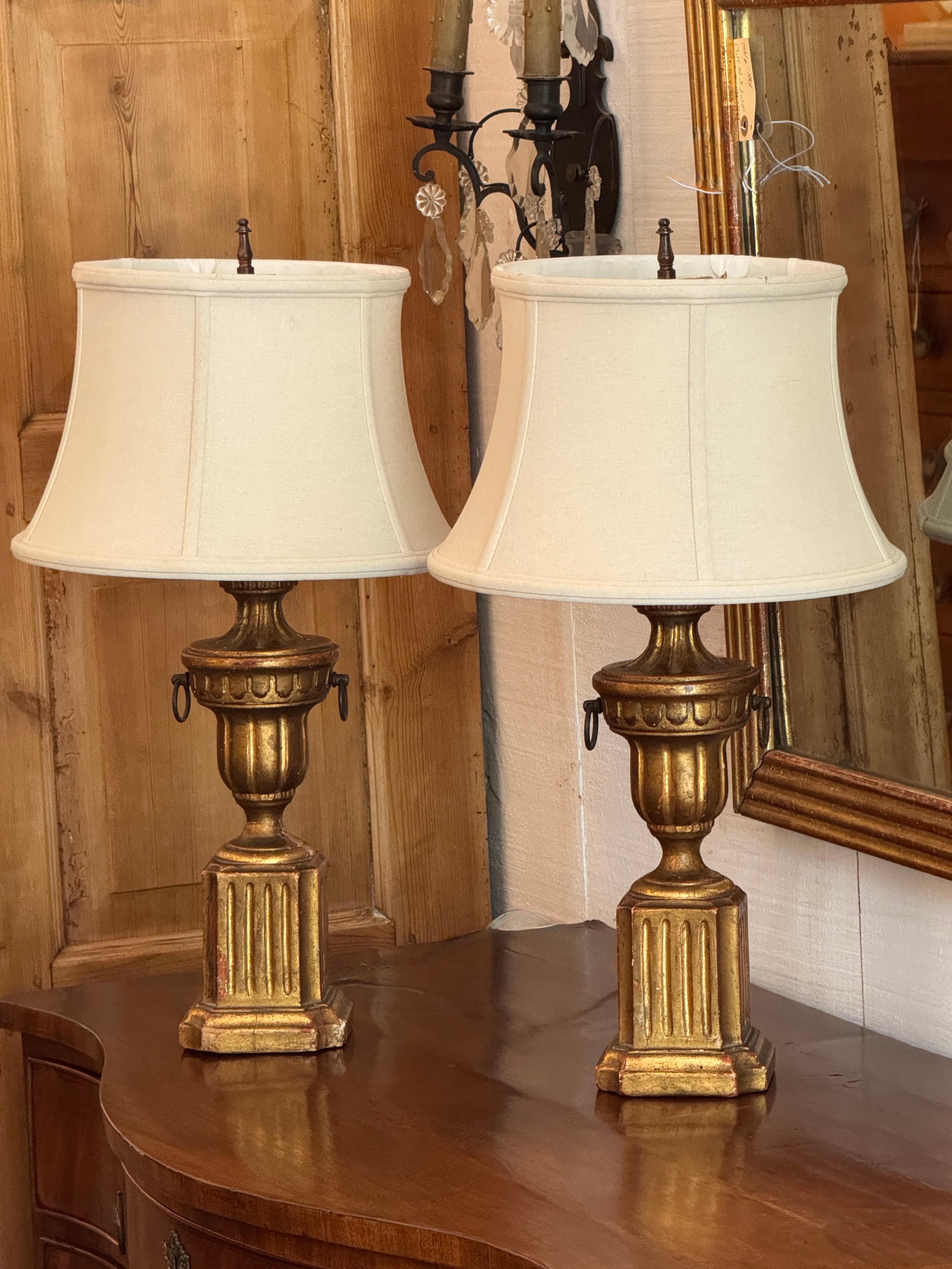 Classic pair of Borghese-style table lamps with carved and gilded urn-form bases. The fluted columns and block plinths evoke neoclassical design, with timeworn gilding adding warmth and age. Subtle ring handles and cream shades complete the look. A