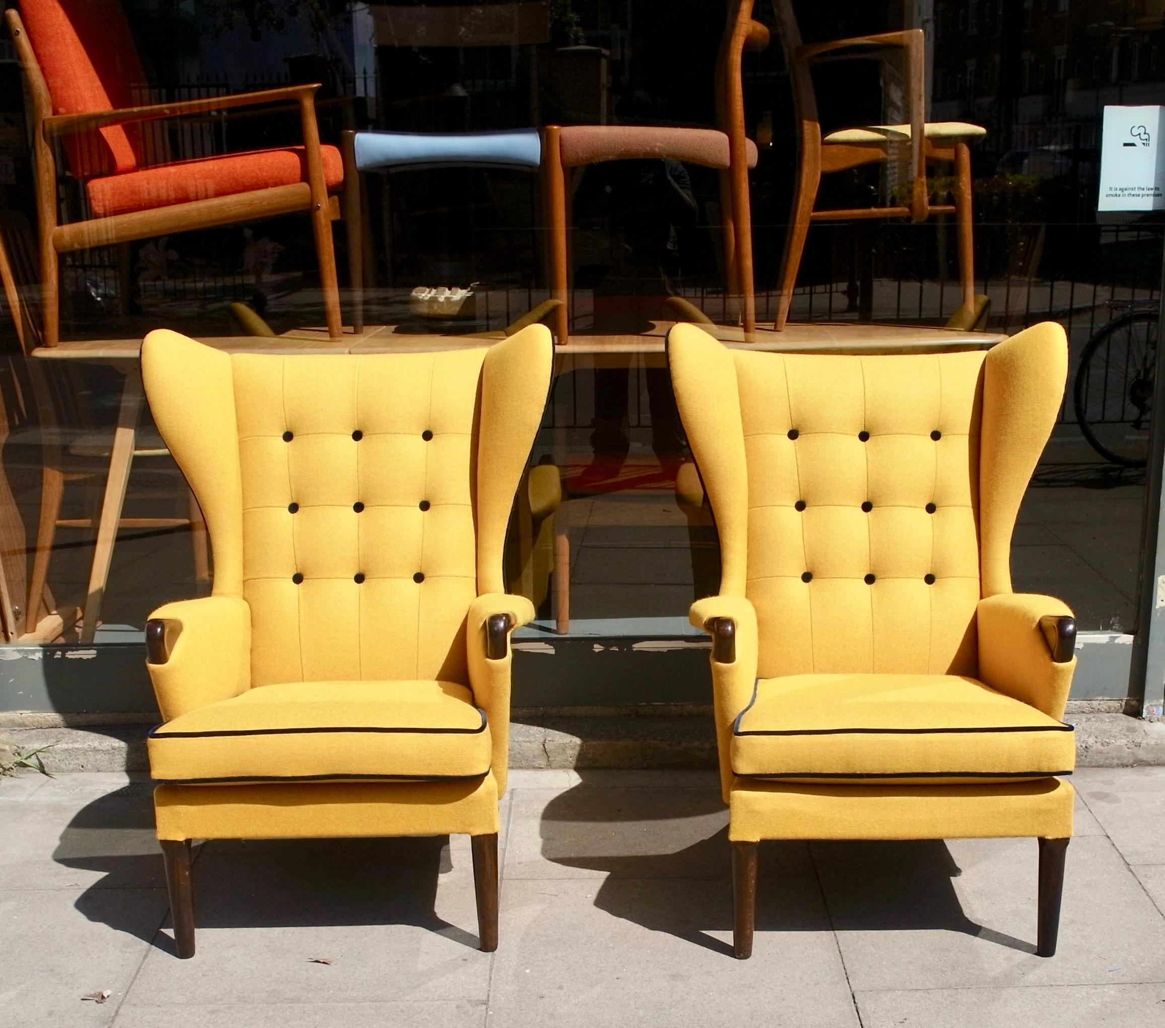 Une paire de fauteuils anglais à dossier ailé boutonné, très confortables et élégants, datant des années 1950. Nouvellement remeublée dans un textile de qualité de couleur jaune, avec un passepoil et des boutons en velours marine, qui accentue le