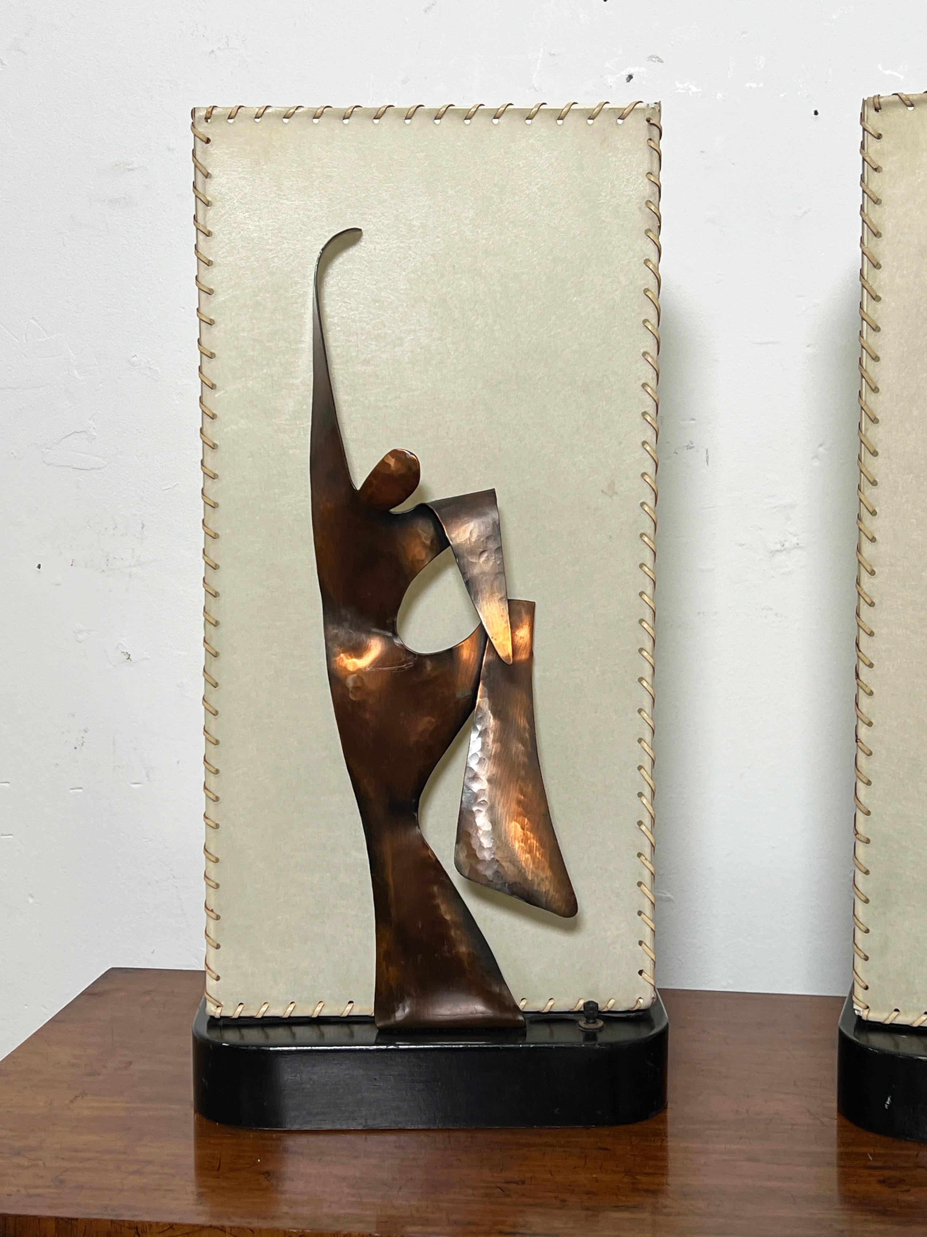 Paire de lampes de table signées Heifetz avec des figures sculpturales de danse moderne en silhouette de cuivre, avec des abat-jours en parchemin lacés d'origine sur des montures ébonisées, vers les années 1950.