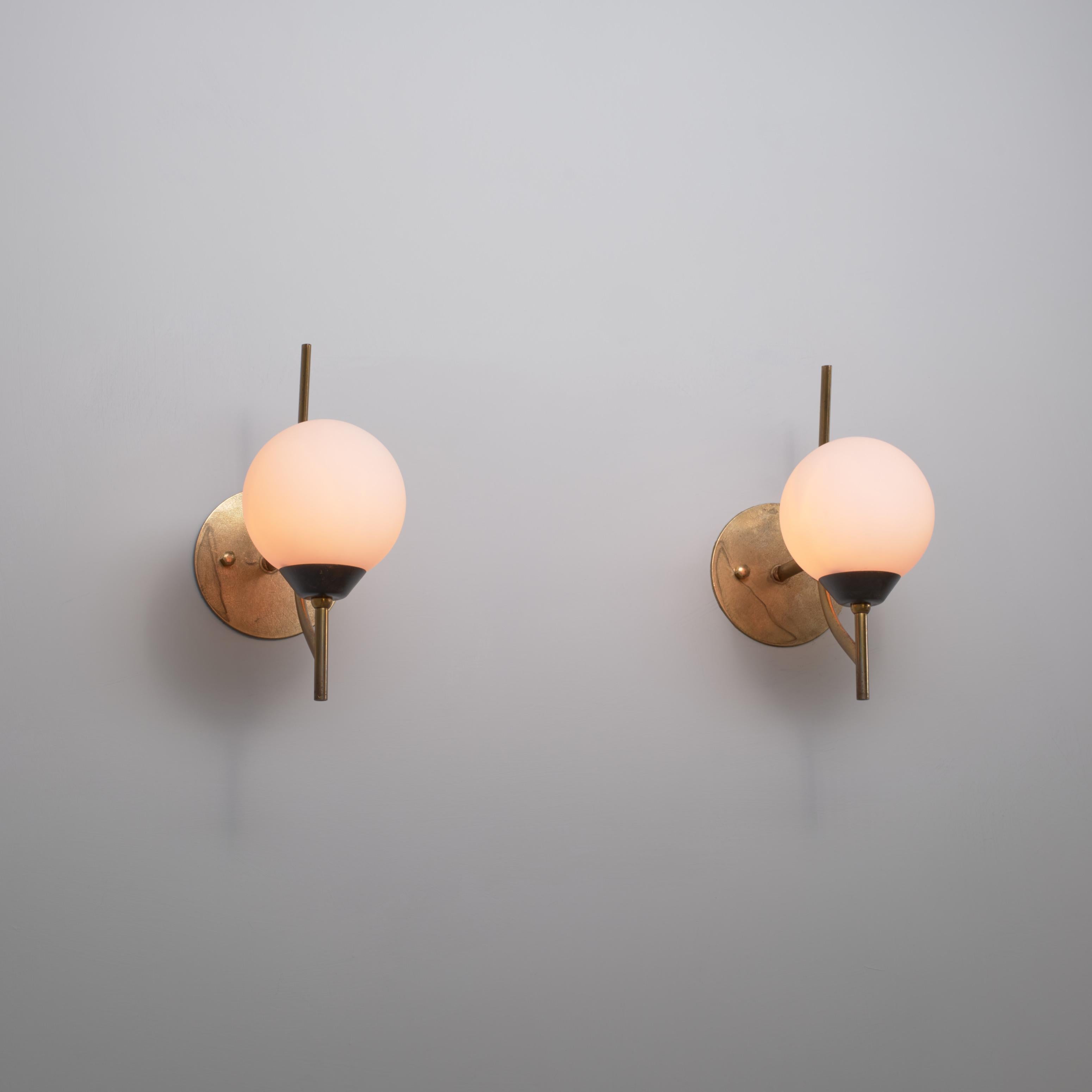 Pair of Italian wall sconces from the 1950s, each composed of a curved brass arm extending from a circular wall plate (12 cm diameter) to support a spherical opaline glass diffuser. The brass surfaces retain pleasing oxidation marks from age, while