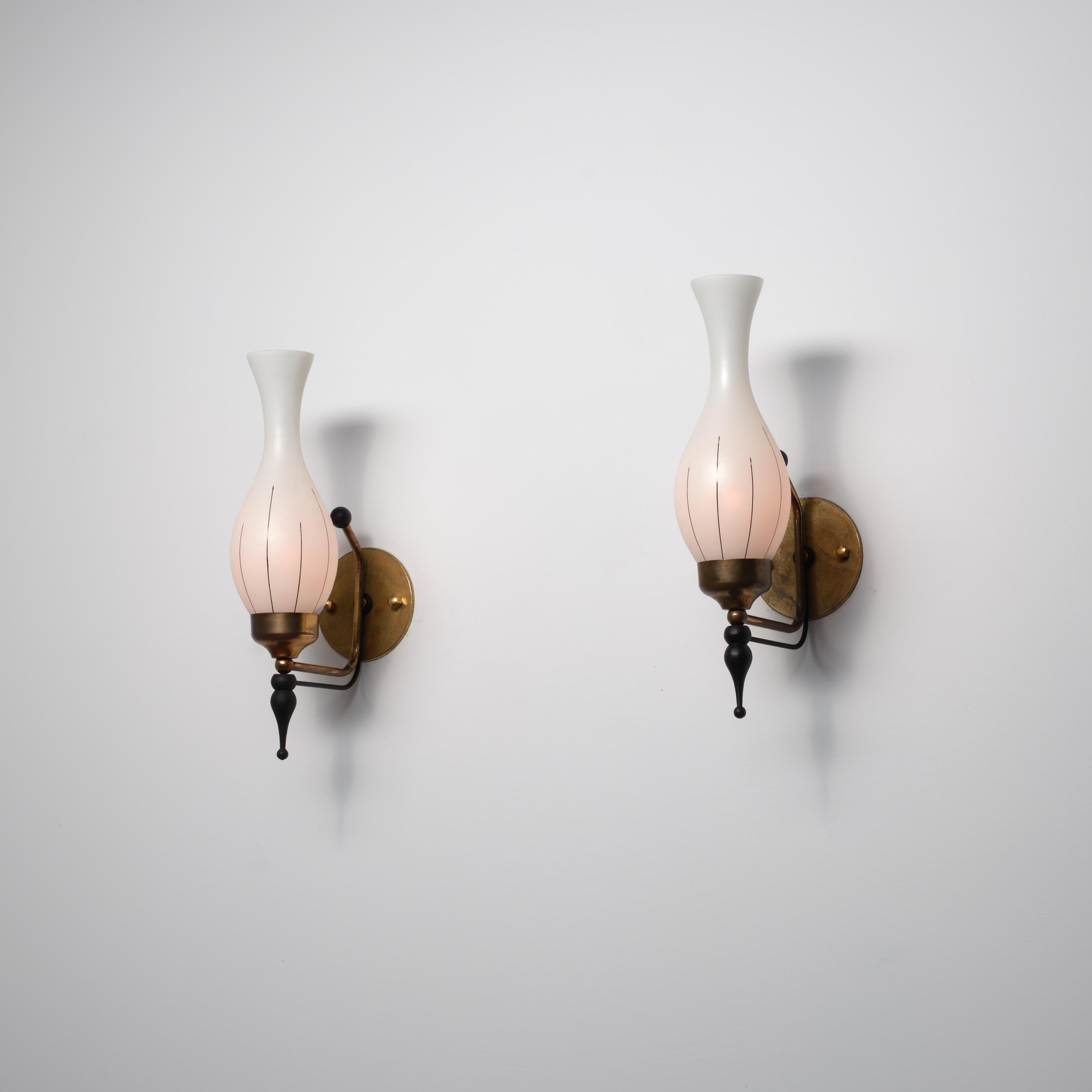 A pair of Italian wall sconces from the 1950s combining brass, black-lacquered metal and hand-blown opaline glass. The slender multi-material structure presents a playful modern design: a curved brass arm supports a drop-shaped finial in