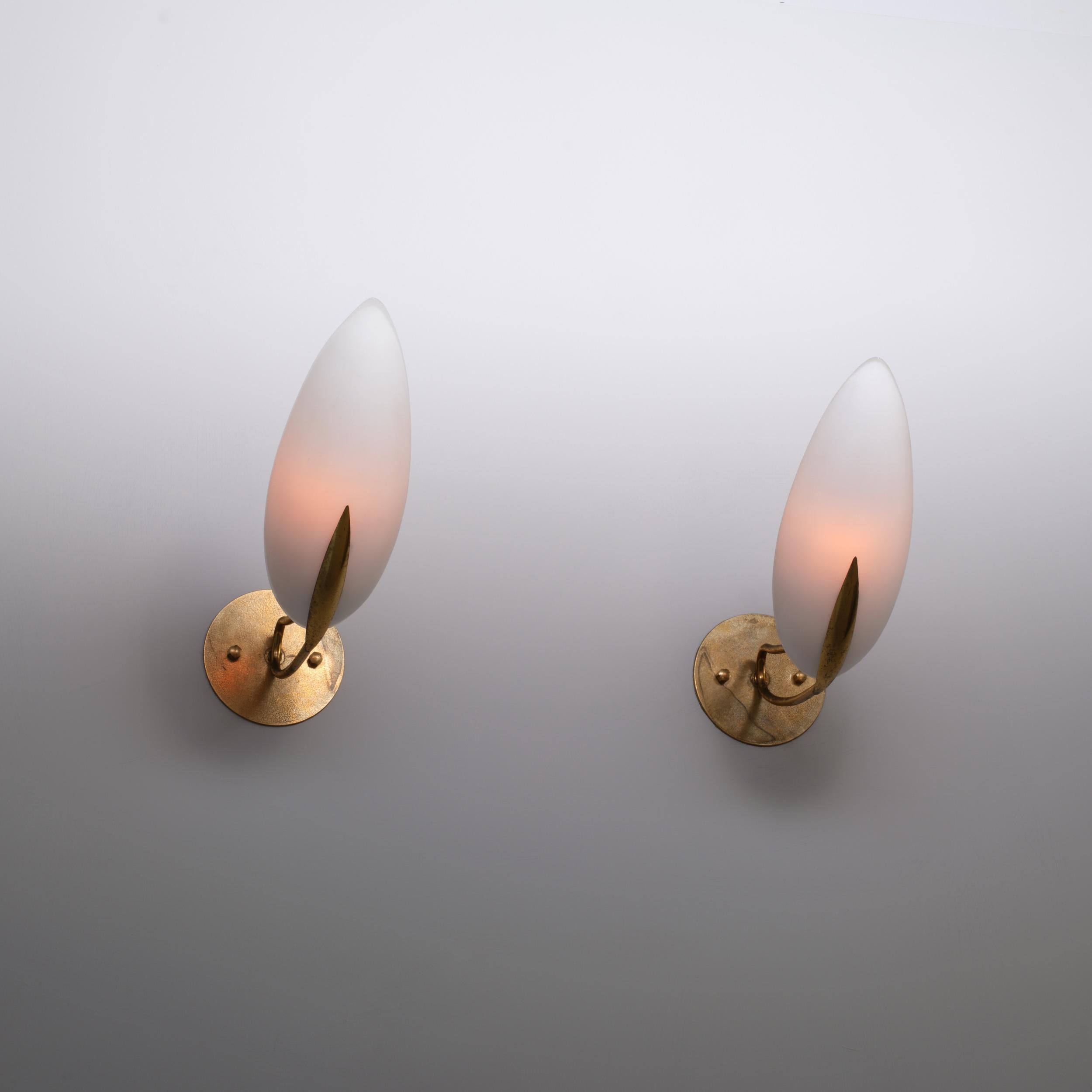 Coppia di applique italiane degli anni '50, ciascuna caratterizzata da una struttura in ottone con due bracci curvi che si estendono da una piastra circolare (diametro 12 cm) per sostenere diffusori in vetro opalino a forma di mandorla. Le superfici