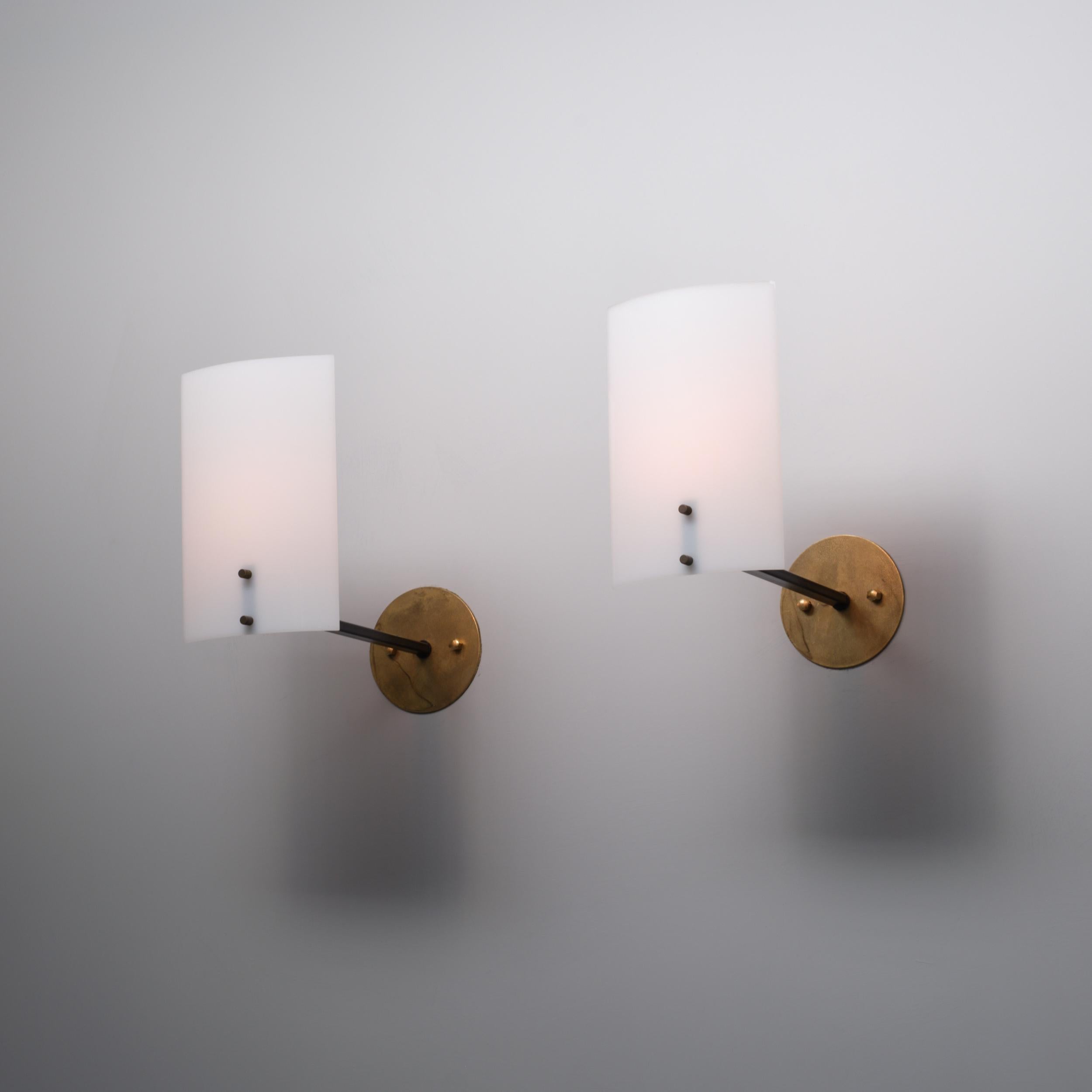 Pair of Italian wall sconces from the 1950s, each composed of a brass wall plate (12 cm diameter) from which a slender black lacquered steel arm extends to support a curved white diffuser in opaline methacrylate . The brass elements show appealing
