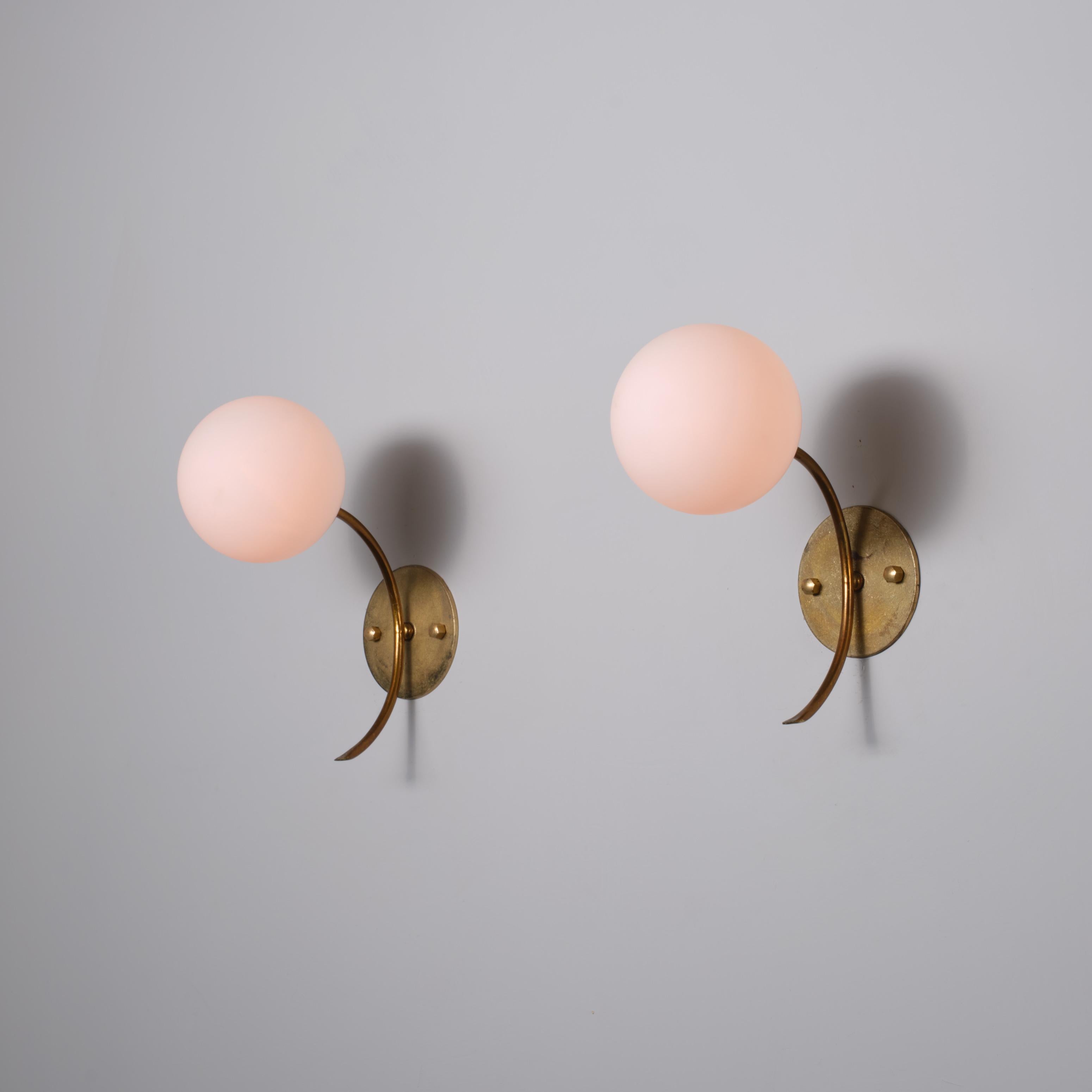 Pair of Italian wall sconces from the 1950s, each featuring a curved brass arm extending from a circular wall plate to support a spherical opaline glass diffuser. The brass surfaces display attractive oxidation patterns developed over time,