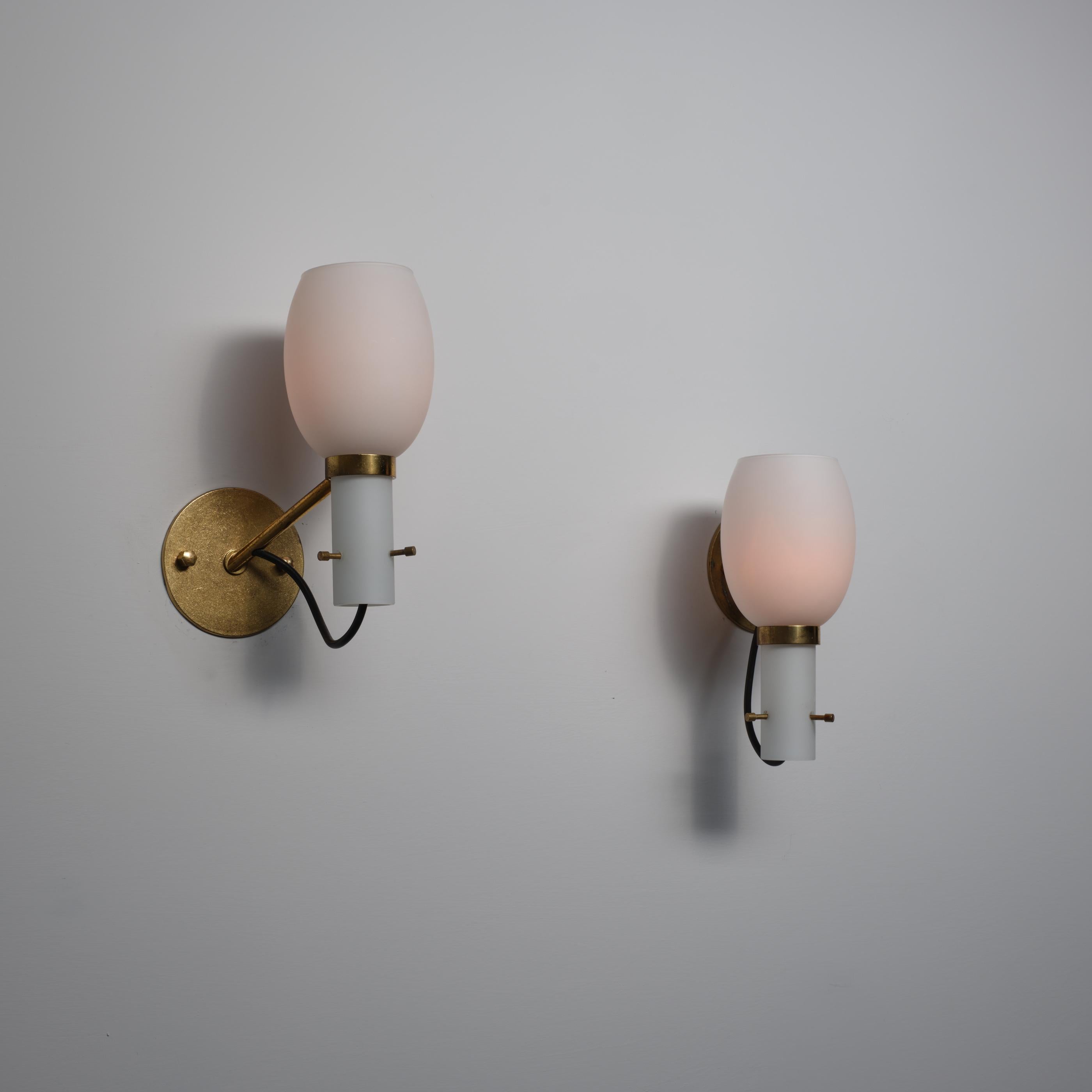 Pair of Italian wall sconces from the 1950s featuring a minimal yet refined composition in brass and opaline glass. Each light presents a cylindrical opaline glass holder supporting a softly rounded shade that diffuses a warm glow when illuminated.