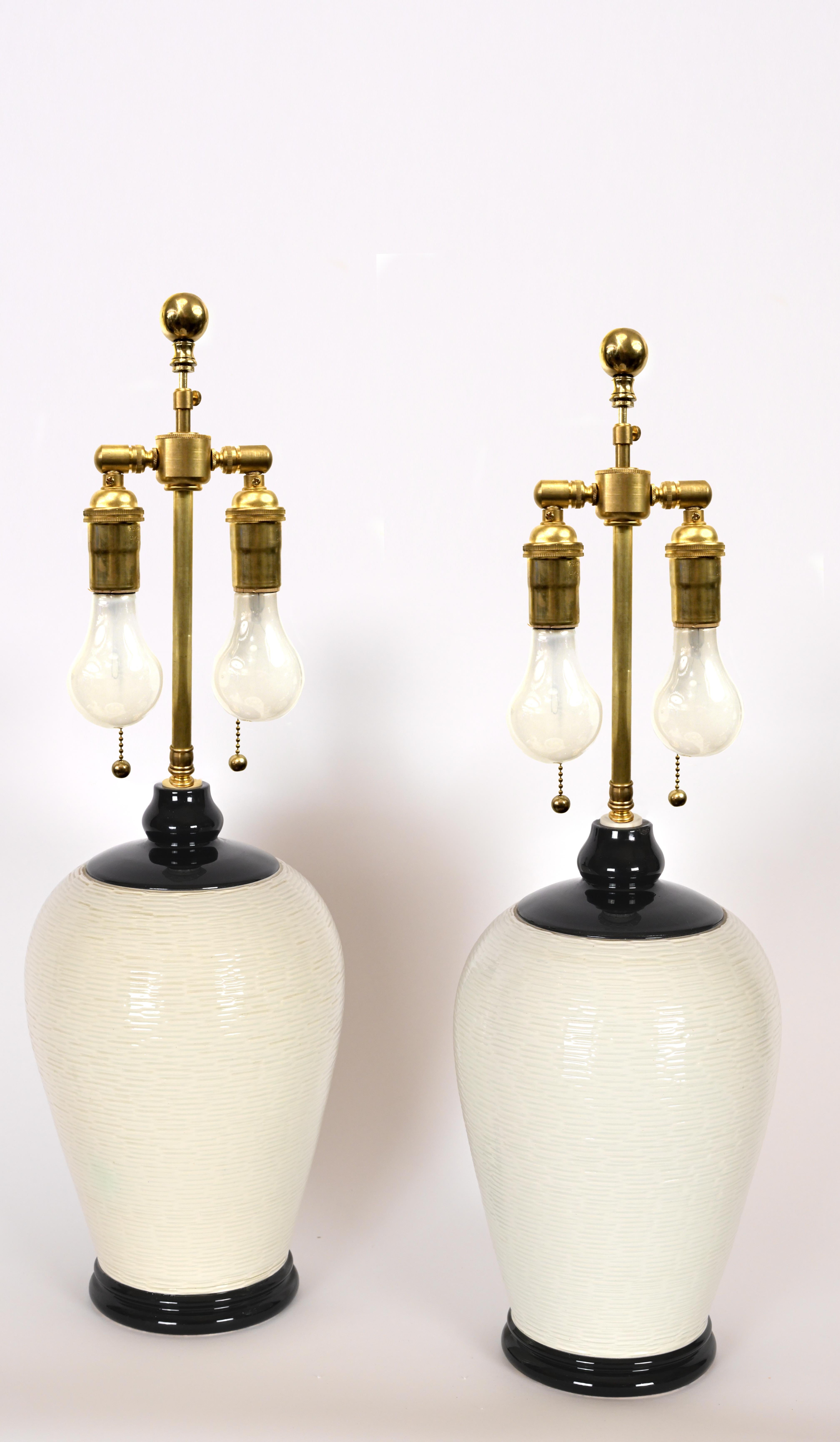 A pair of white ceramic lamps made by Nordiska Kompaniet having black accented top and bottom with ridging along the sides. Started in 1890, Nordiska Kompaniet is a luxury department store company that has collaborated with famous designers and