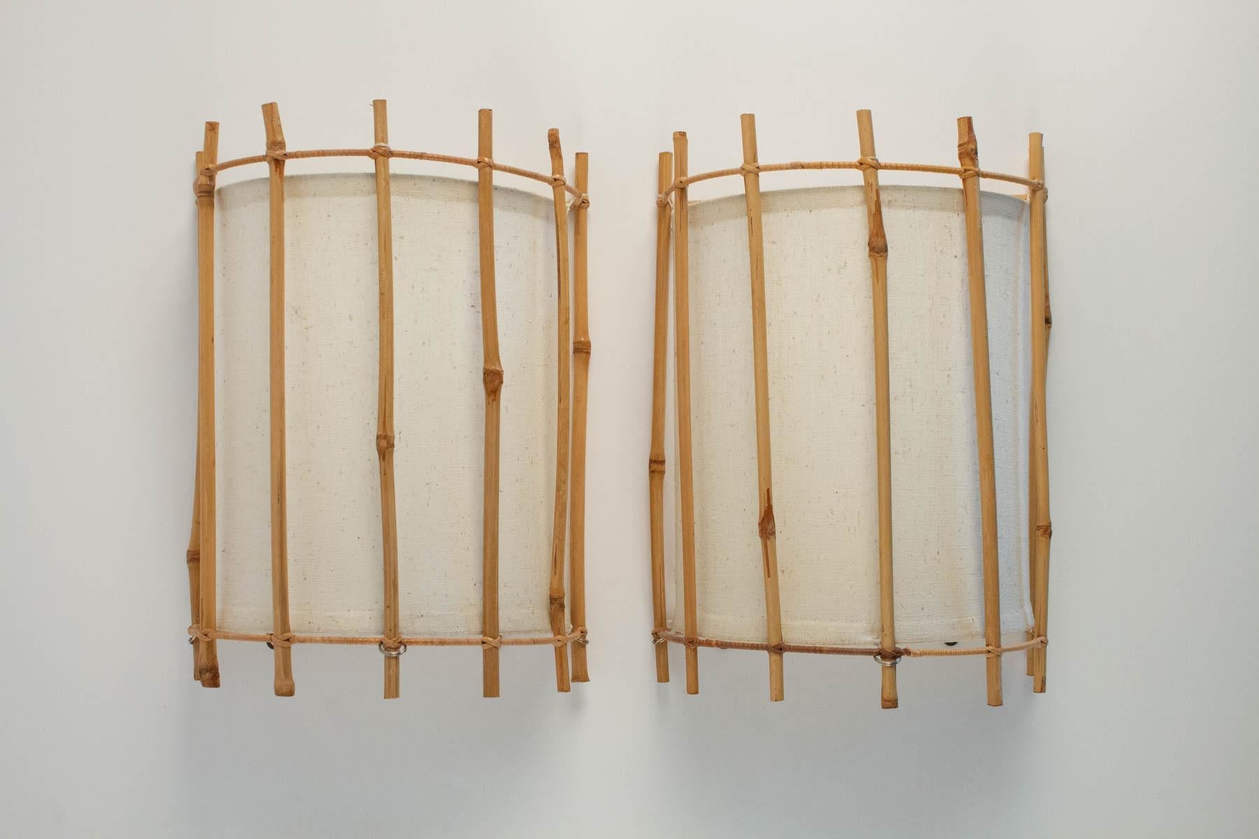 Pair of 1950s rattan sconces by Louis Sognot. Fine vertical rattan wands thighed with two horizontally curved rattan wands. Off-white cotton lampshades. One lighted arm per sconce.