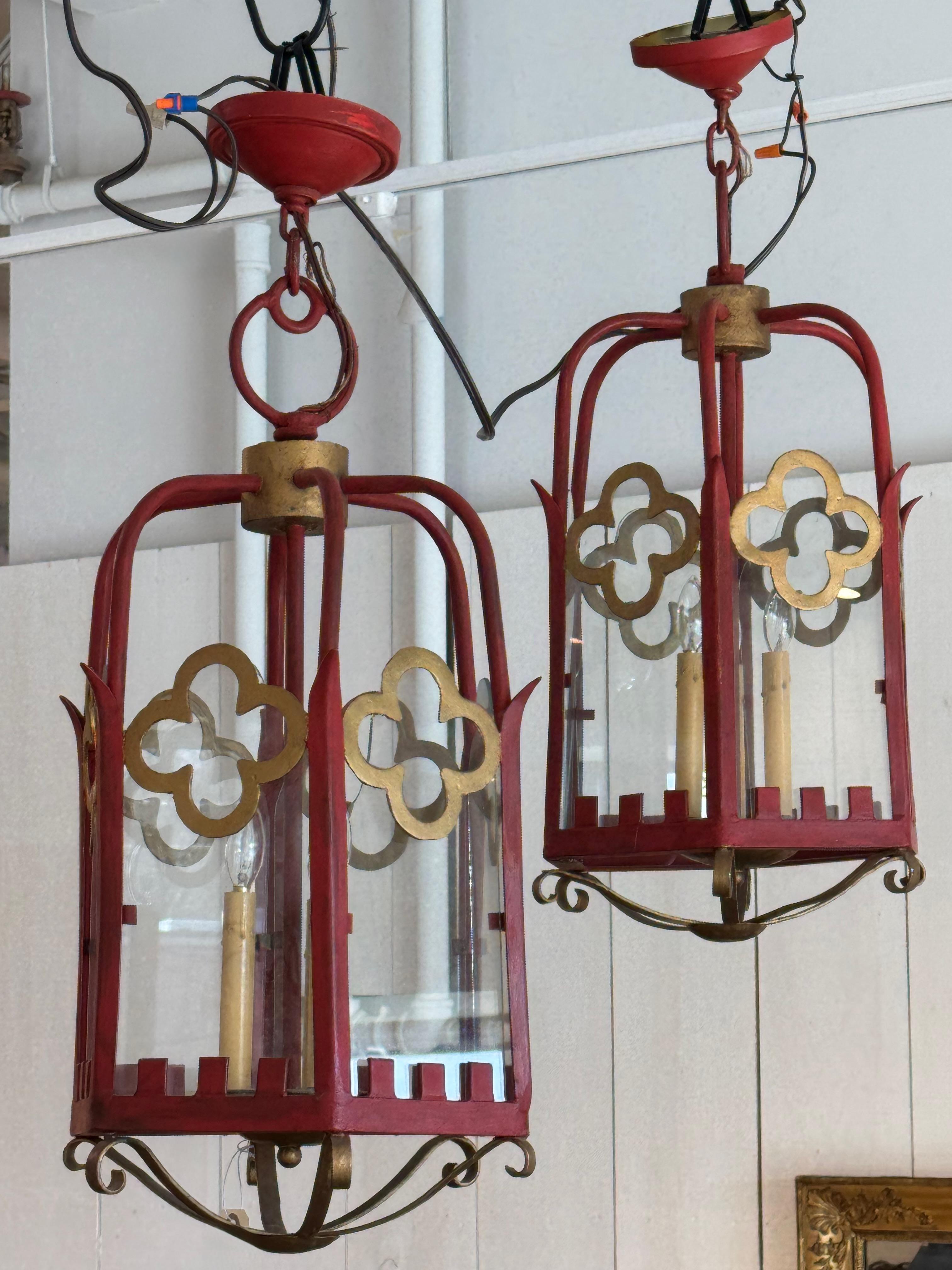 Striking pair of red-painted metal lanterns with a Gothic-inspired design. Featuring gold quatrefoil accents and clear glass panels, these lanterns combine decorative charm with functional lighting. The curved metal framework and scrolled base