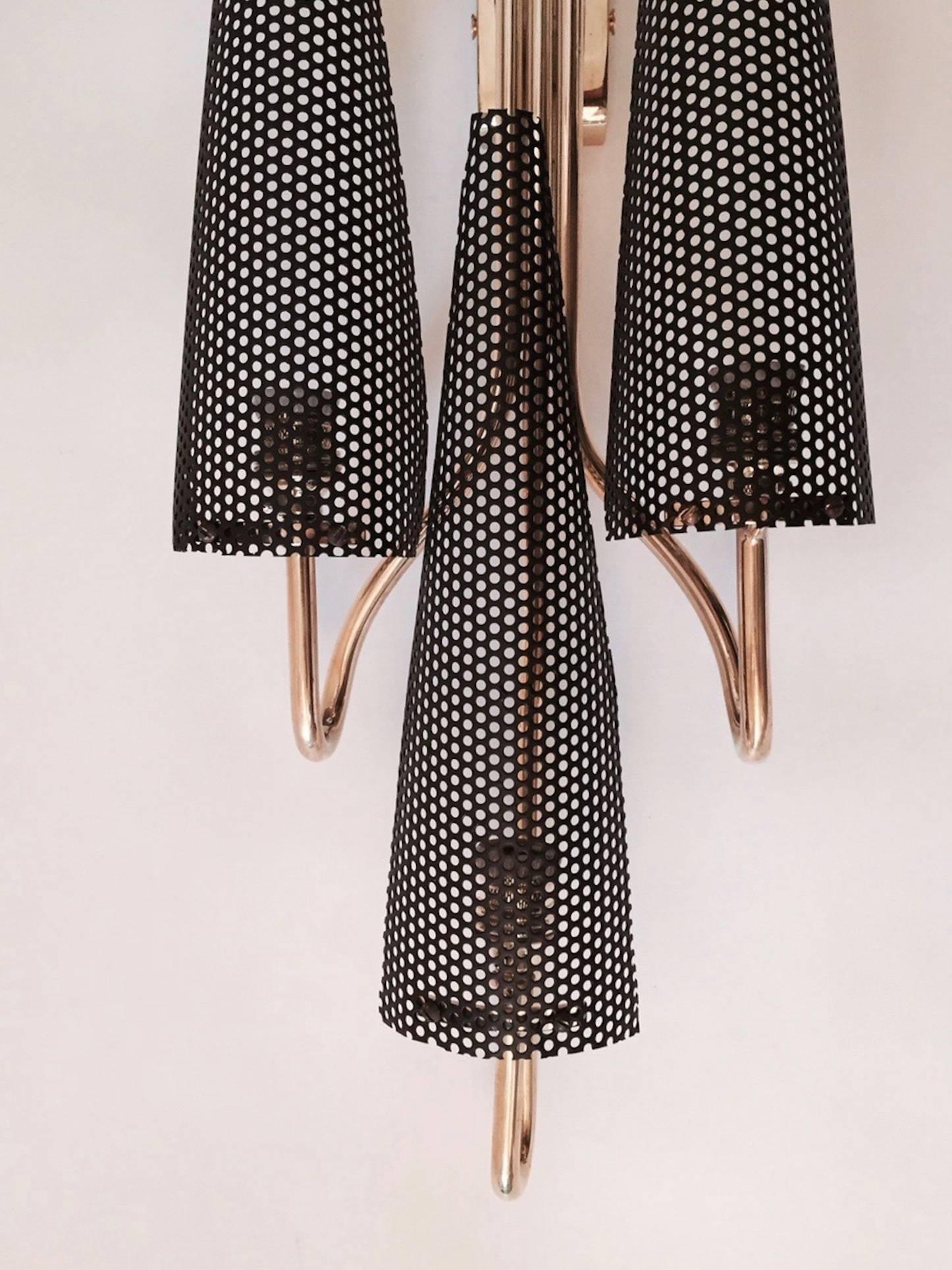 Pair of 1950s sconces attributed to Jacques Biny.
The sconces are composed of three gilt brass rods splitter on the base. 
They are highlighted by black perforated steel sheet semicircular lampshades. Three lighted arms per sconce.