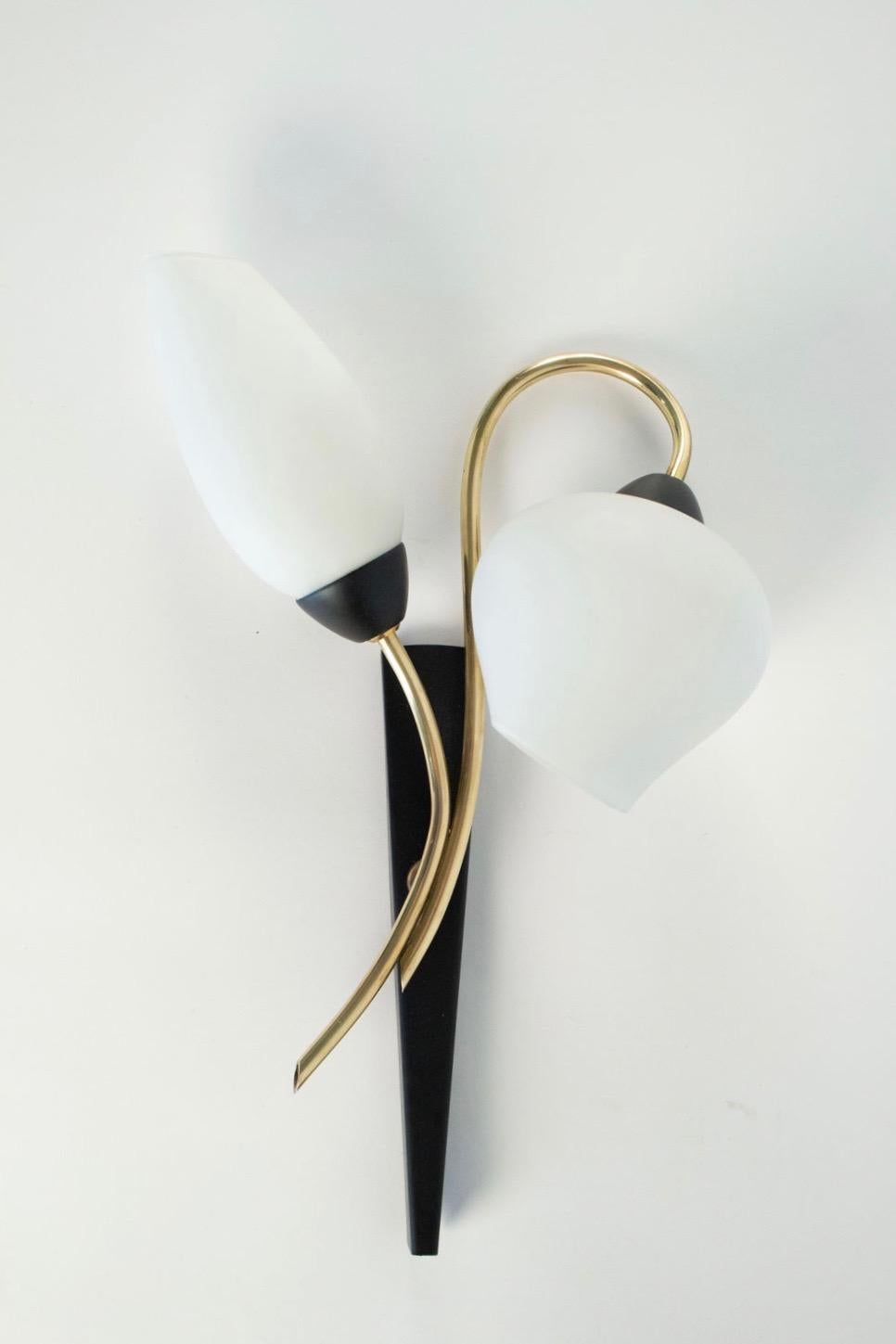 Pair of 1950s sconces by Maison Lunel.
Each sconce consisting of two gilt brass arms of a stylized flower shape surmounted by truncated ovoid opaline glass suggesting the flower button.
The trapeze shape black metal base underlines the piece.
Two