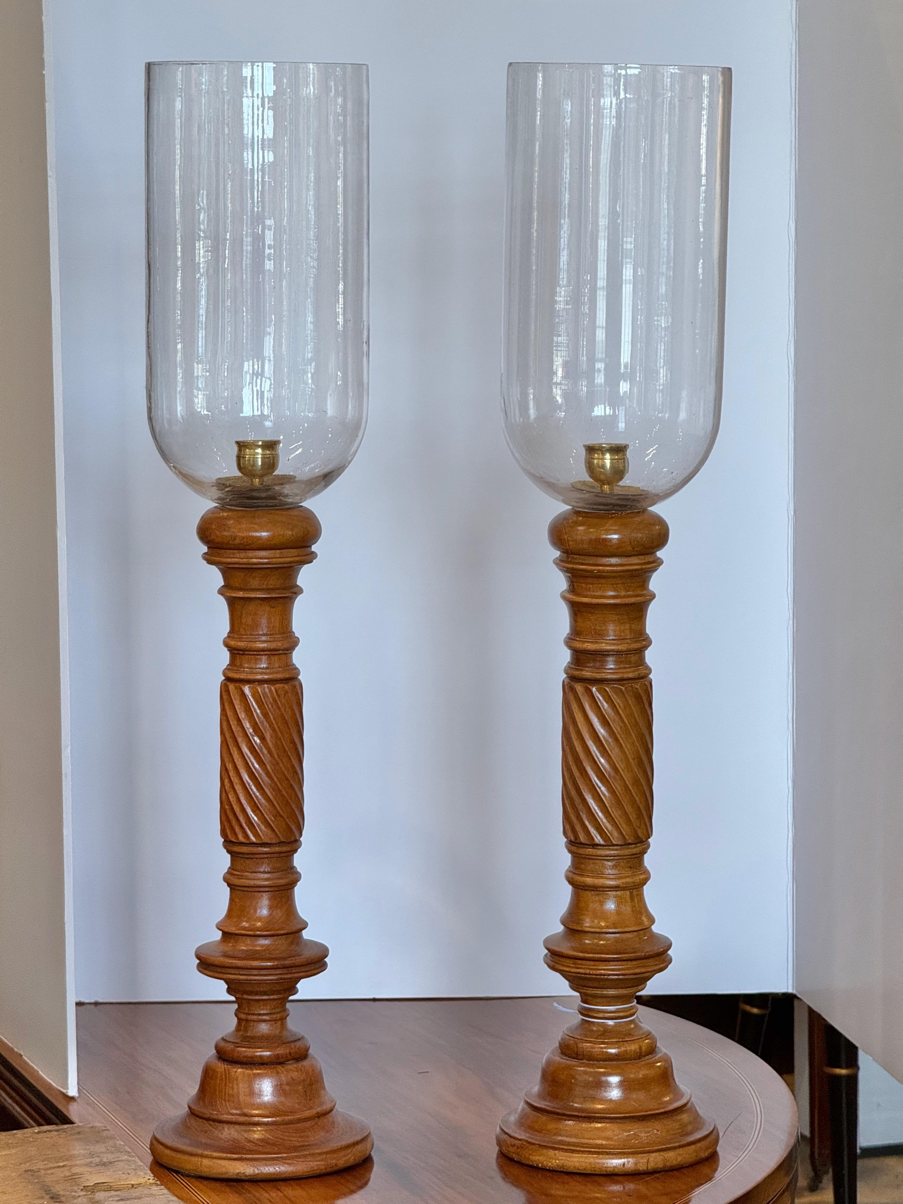 Elegant pair of hurricane lamps featuring hand-blown glass shades with a subtle waviness, set on turned and carved wooden bases. The tall, gracefully proportioned columns showcase a rich wood grain with decorative spiral carving. Brass candle