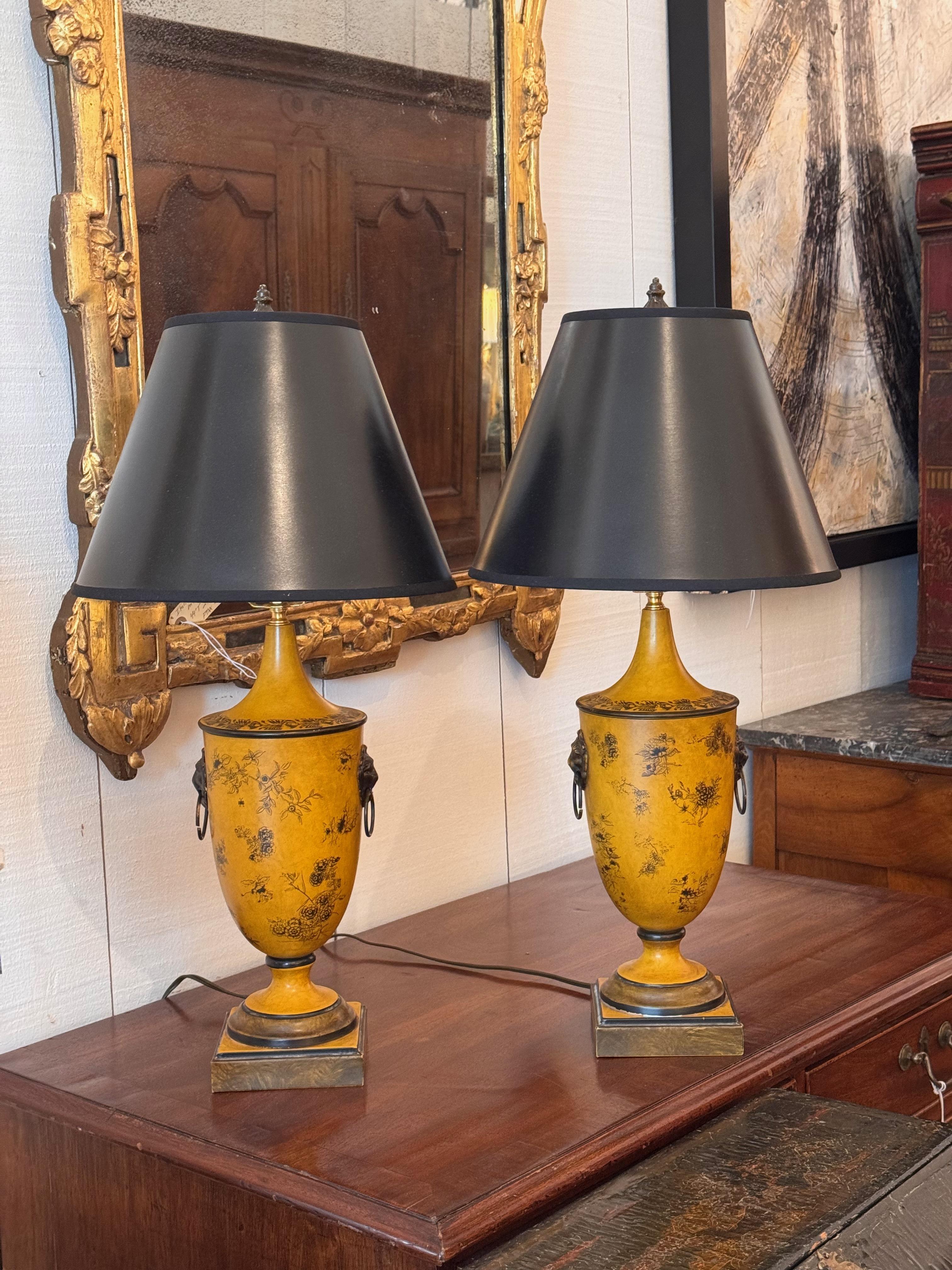 Pair of urn-form tole lamps in a bold mustard yellow, decorated with fine black floral detailing and mounted with cast lion mask ring handles on each side. Set on squared plinth bases with black-painted accents and fitted with black empire shades. A
