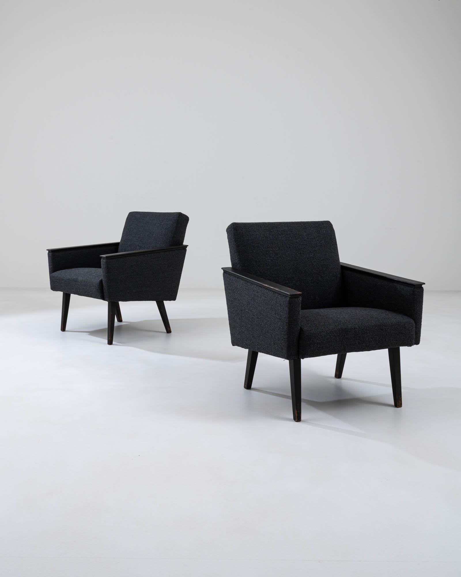A pair of upholstered wooden armchairs from France, produced circa 1960. A smart set in a deep black, featuring black patinated wooden armrests and legs. Chic slants create a sloping seat back and trapezoidal side panels in this conversational pair.