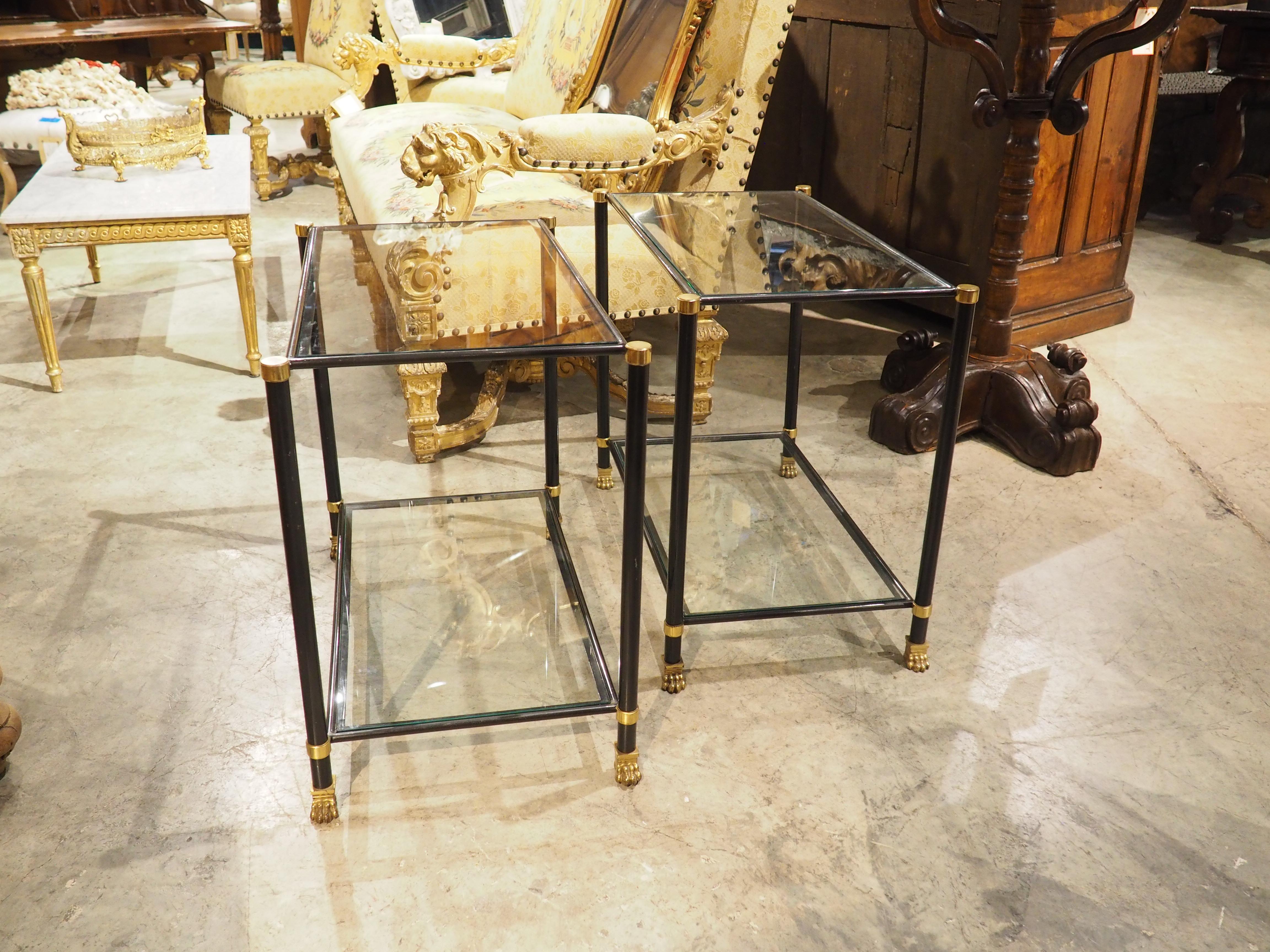 Esta pareja de mesas auxiliares francesas, de los años sesenta, presenta una disposición rectangular de cristal en dos niveles. Ambos cristales se apoyan en un marco metálico rectilíneo pintado de negro, con todos los bordes coronados por