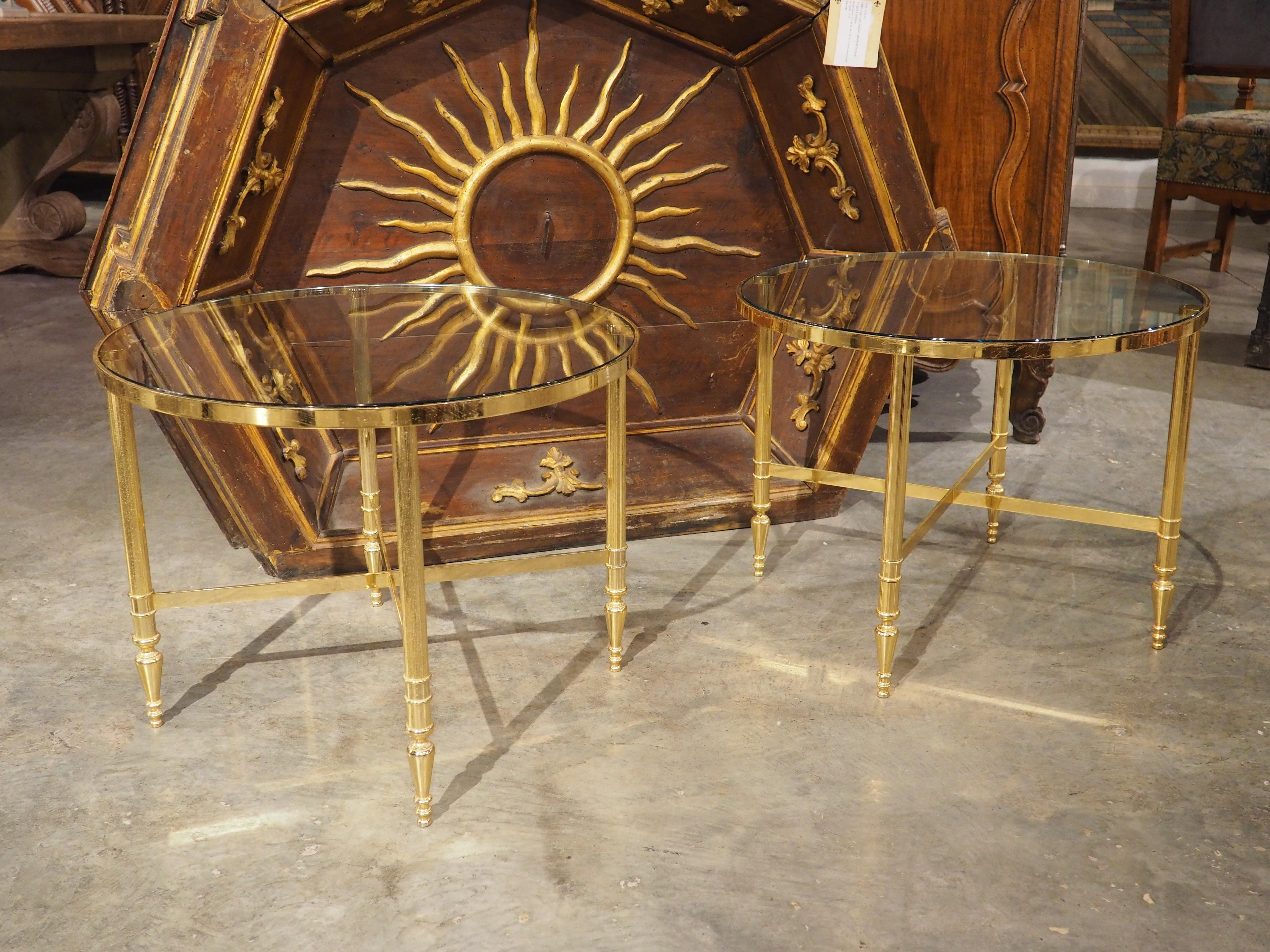 A lovely pair of French side tables, dating to the 1970s, constructed with brass bases and 3/8 inch thick inset circular glass.  The brass has a vibrant gold color, with each of the four slender legs embellished with slender fluting.  An X-stretcher