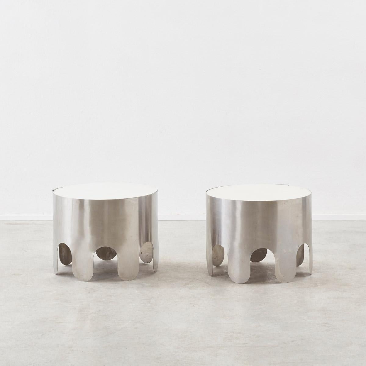 A pair of playful round brushed steel side tables, elegantly balancing soft curves with a more industrial aesthetic. The gentle undulating bases form feet, giving the illusion of waves. The cylindrical metal bases hold off-white resin tabletops,