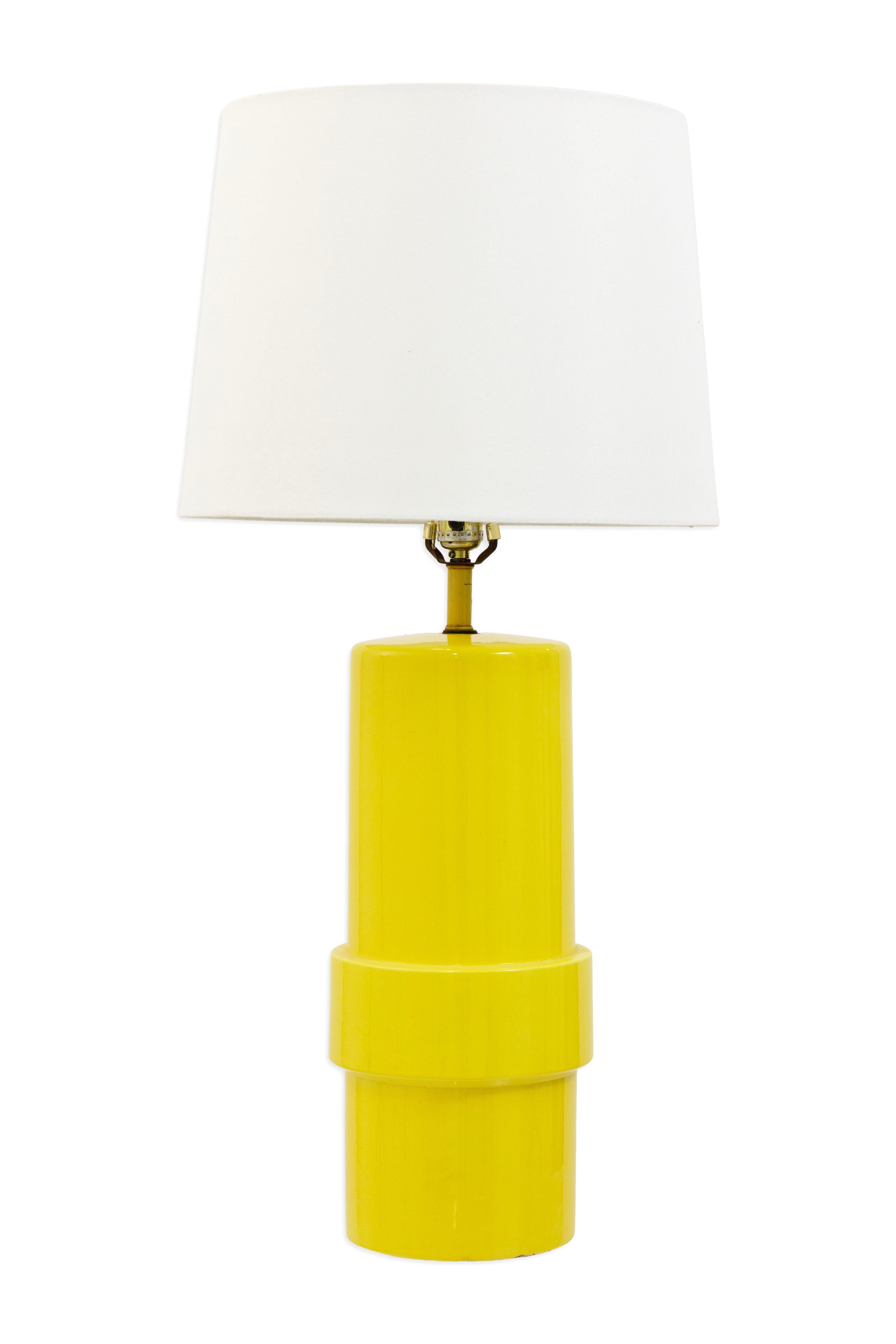 This pair of 1970s ceramic table lamps features tall cylindrical bases finished in a glossy yellow glaze. Each lamp has a simple stacked form that adds a bit of dimension while maintaining a clean look. Topped with white tapered shades, they provide
