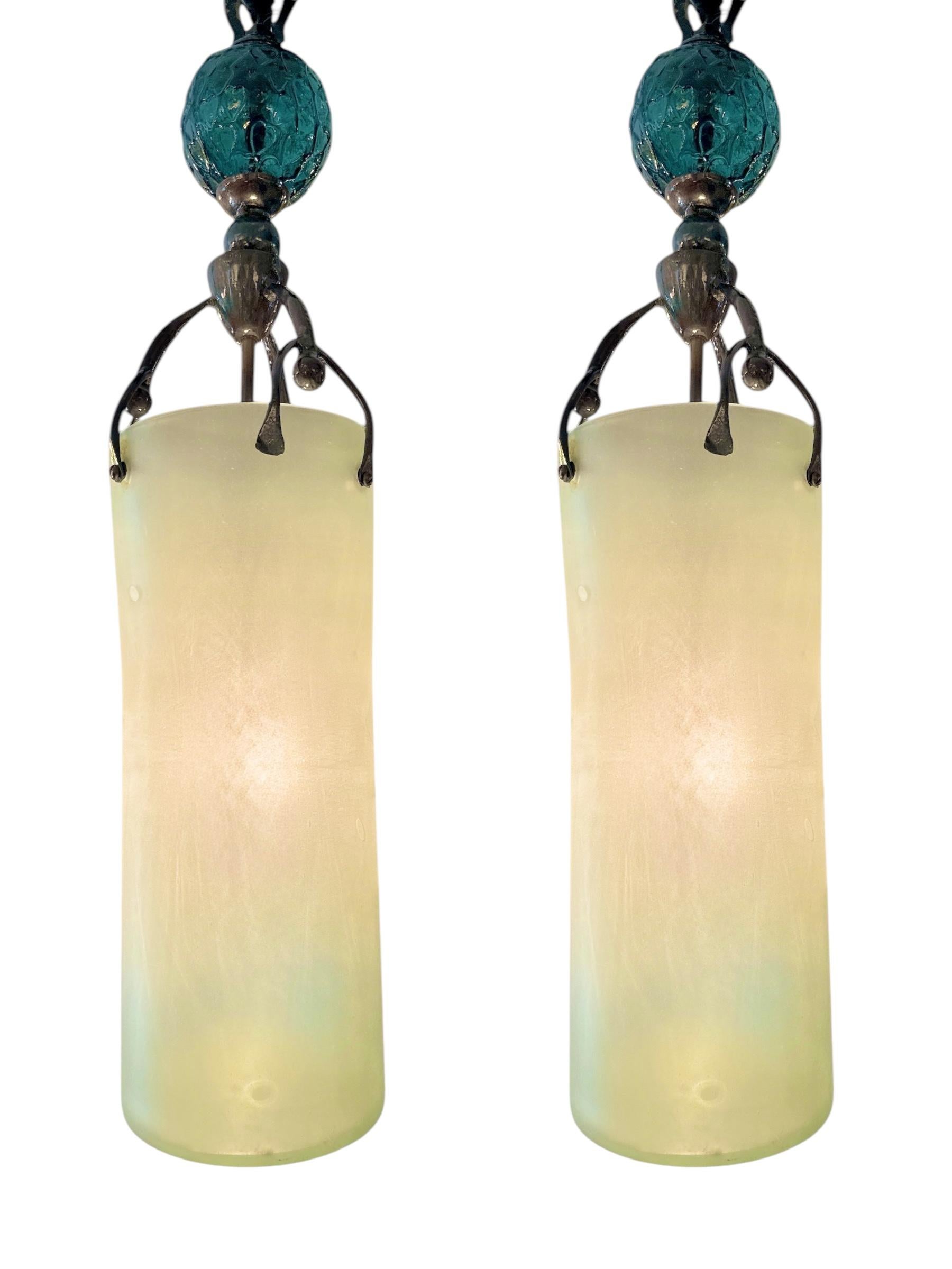 Audace e artistica, questa sorprendente coppia di lampadari brutalisti degli anni '80 è caratterizzata da cilindri in vetro smerigliato soffiato a mano sospesi da bracci scultorei in metallo con patina ossidata. Ogni pezzo è sormontato da una sfera