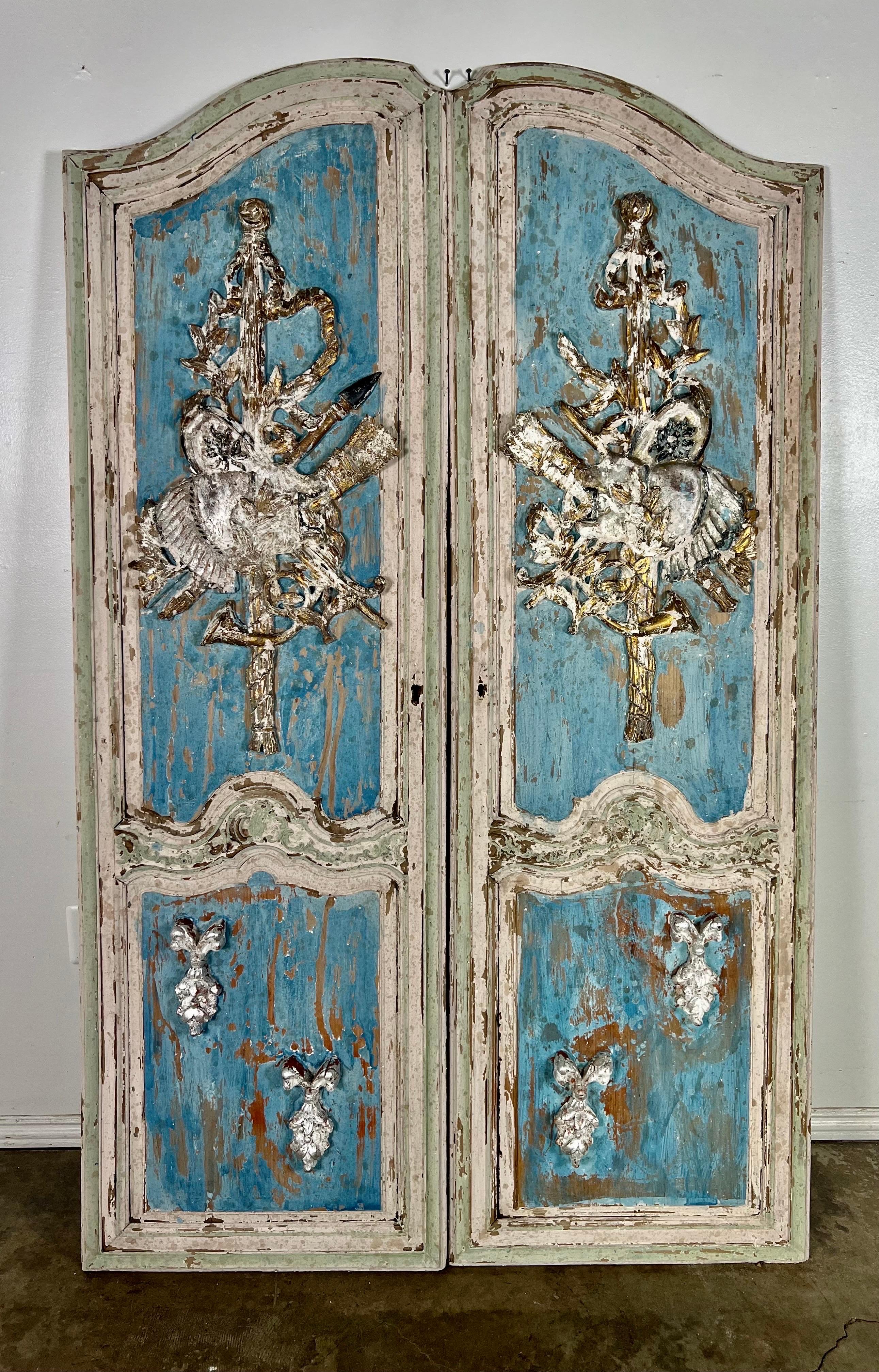 Pair of hand painted antique French doors.  They have beautiful appliqued carvings of musical instruments surrounded by flowers and bows.  They are painted a French blue with accents of white and the natural wood underneath.  The doors are 25