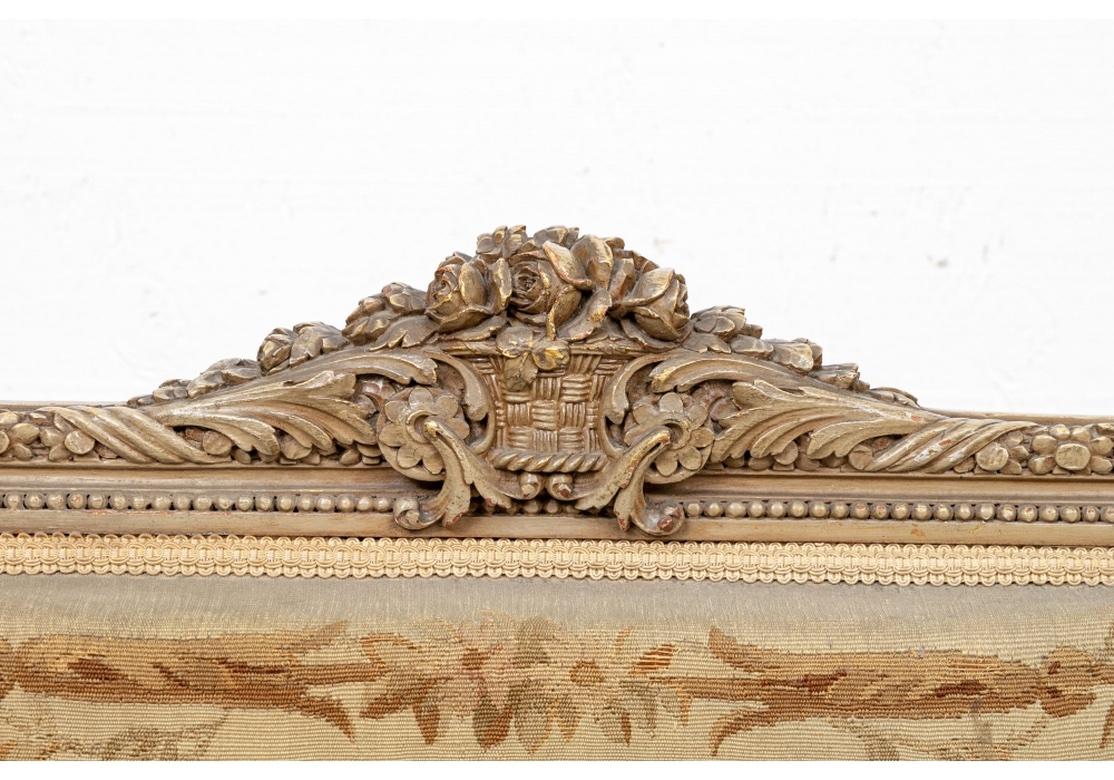 Elaborately carved fauteuils in gray paint with gilt details. The crest rails with floral basket crests and foliate ribbon twist back and seat rail frames. The carved arms with acanthus leaves, raised on fluted legs. Upholstered in worn tapestry in