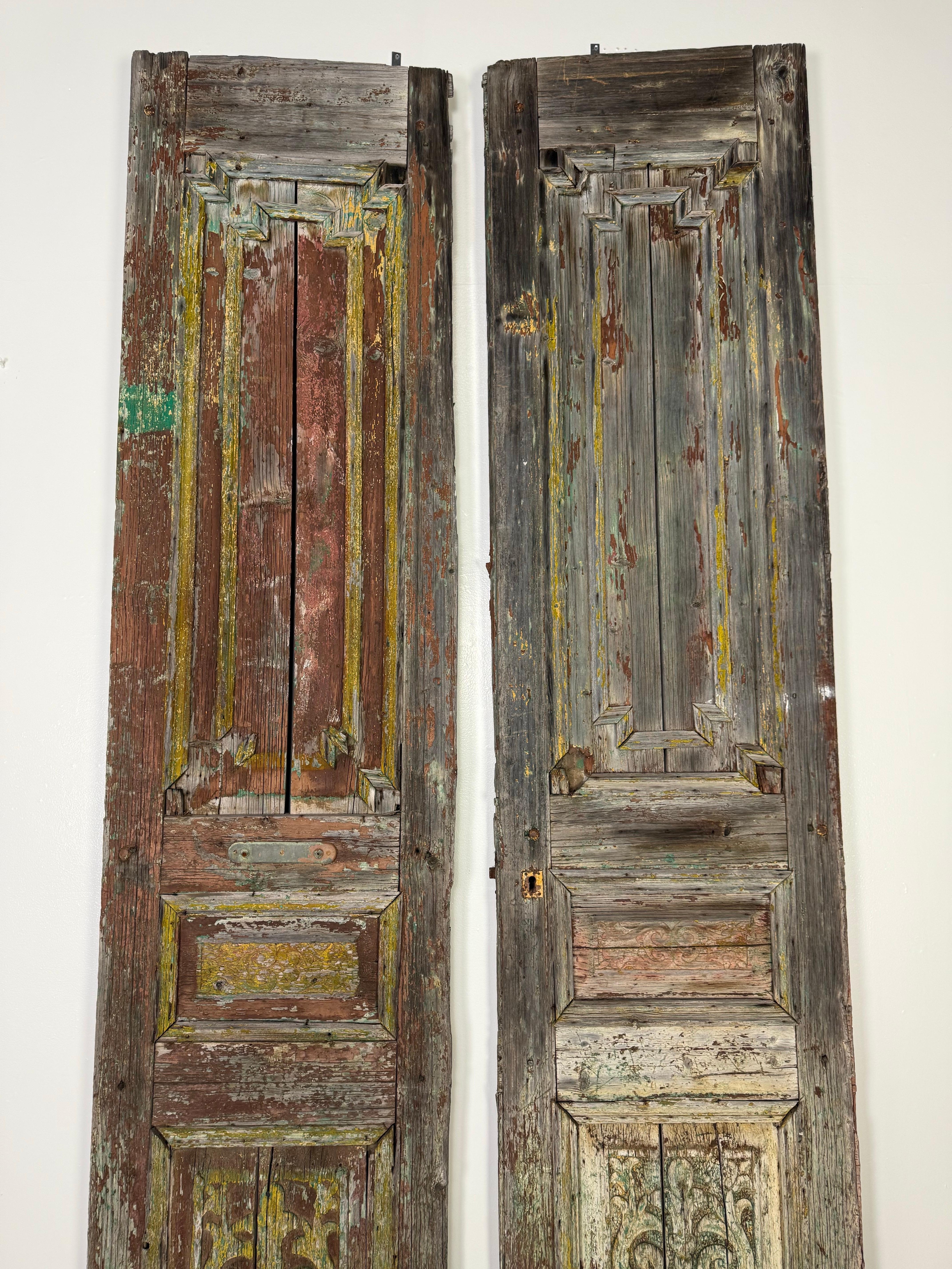 Coppia di porte in legno del XIX secolo dall'aspetto fortemente danneggiato dalle intemperie.  Presentano intricati dettagli intagliati, tra cui pannelli in rilievo e motivi ornamentali, in particolare nelle sezioni inferiori dove sono visibili le