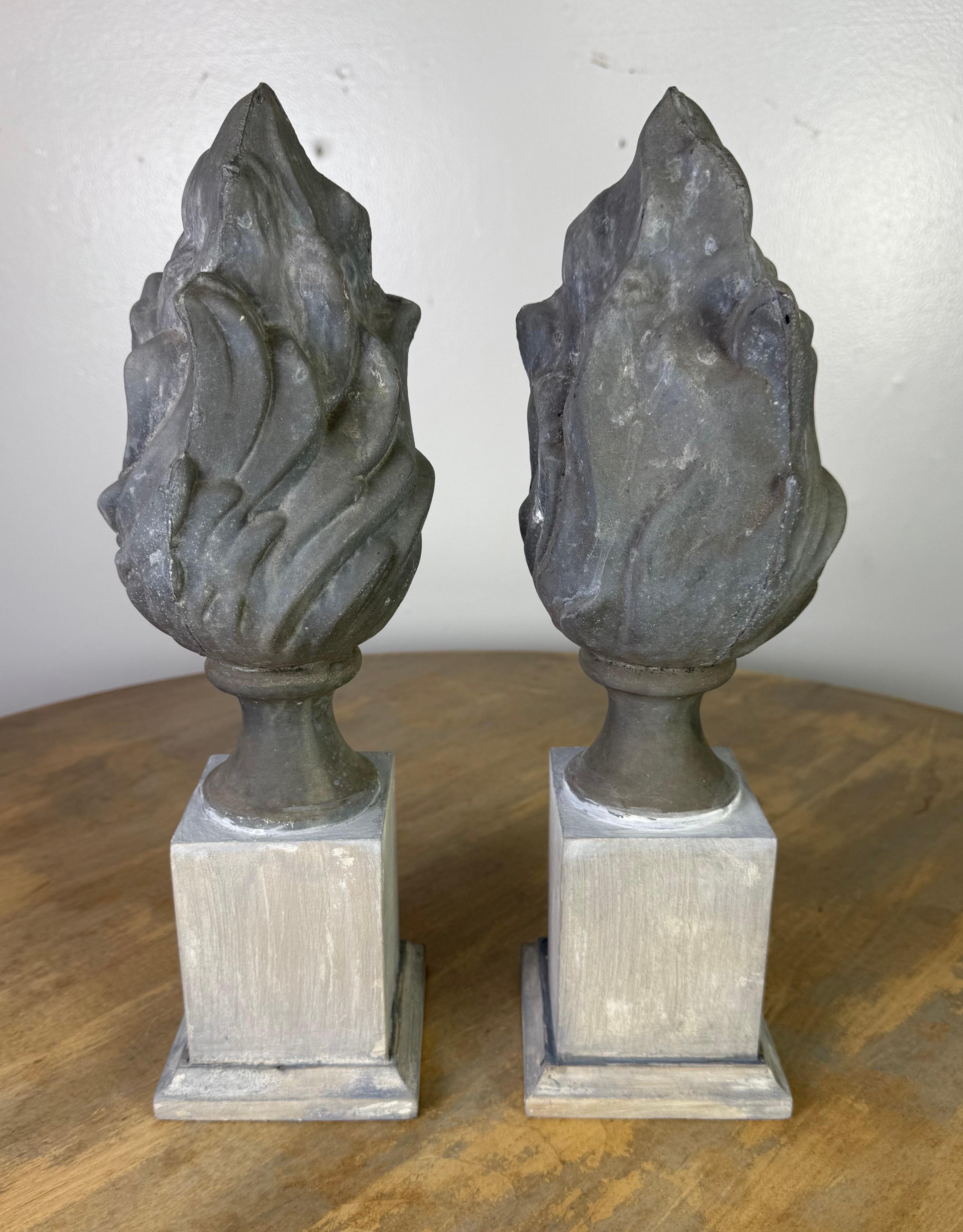 This pair of 19th-century finials features sculptural flame motifs cast in metal, likely zinc, known for its durability and weather resistance. Each flame form rises organically with curling ridges and a tapered peak, giving a sense of movement and