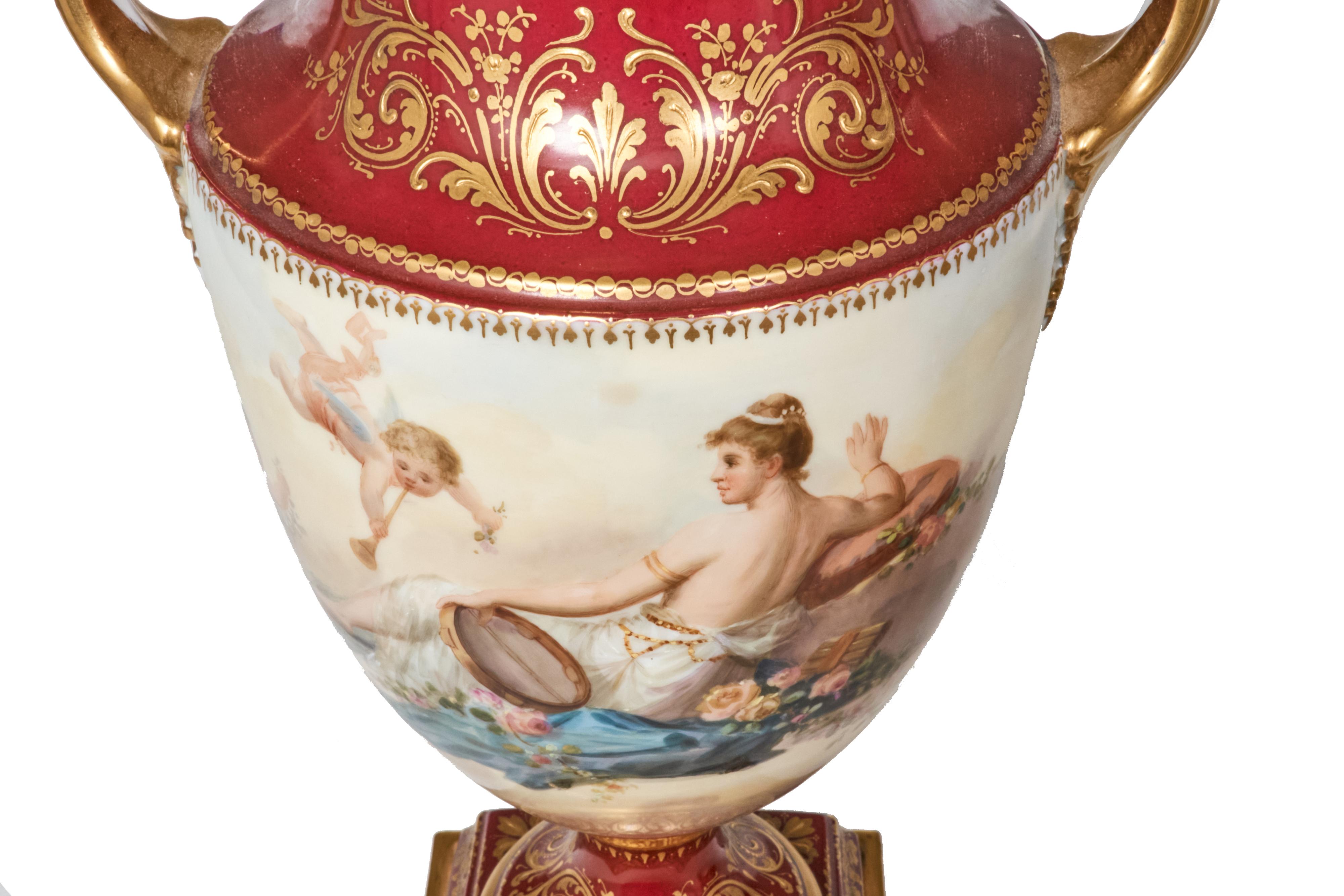 Exquisite pair of hand painted Royal Vienna urns decorated with cherubs and scantily clad women. Rich red is complimented by gold decoration and brass raised bases. Each urn has a matching lid and all pieces are in excellent condition without