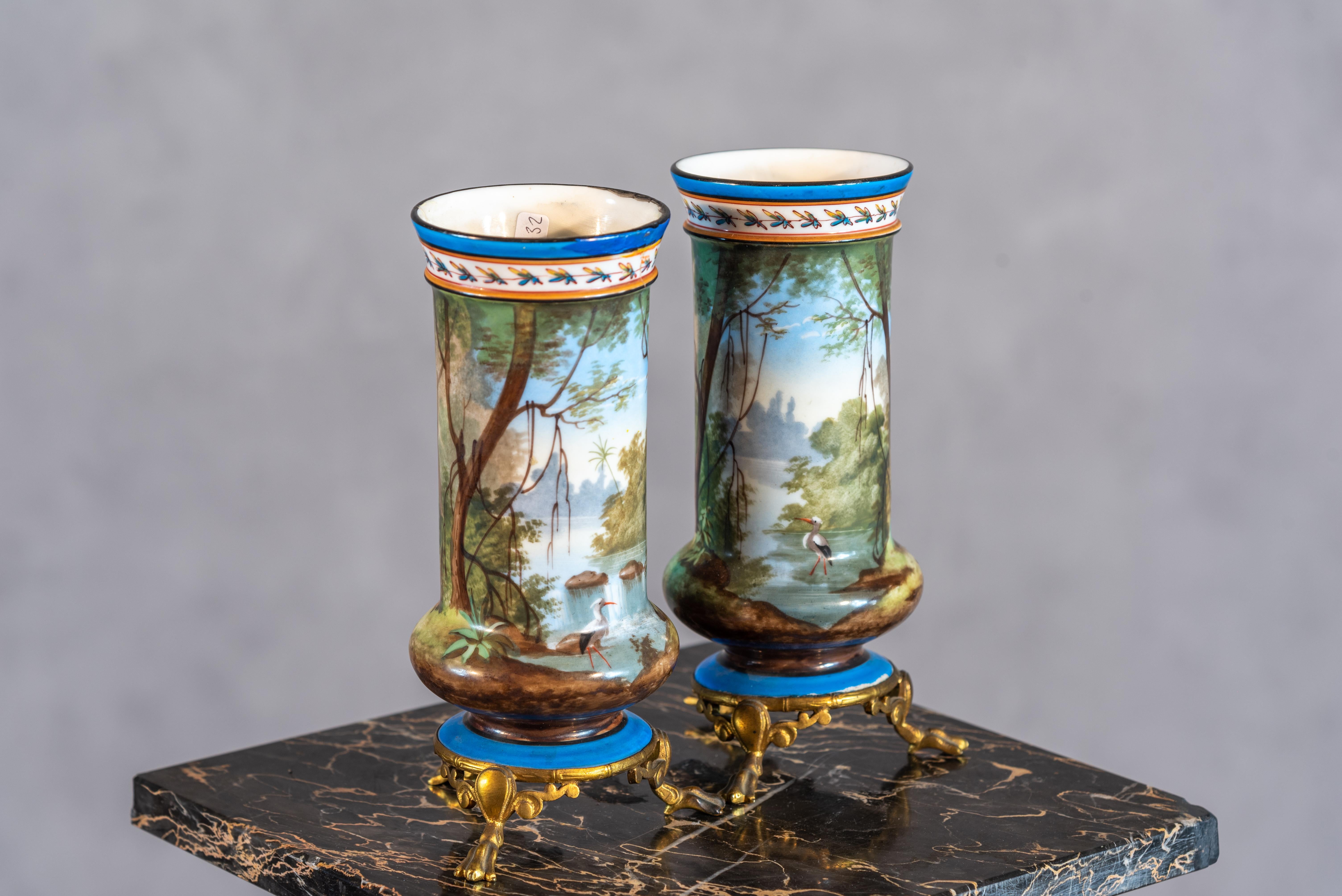 This enchanting pair of 19th-century vases, inspired by the renowned Sevres style, captures a picturesque scene of a tranquil lake framed by a dense forest, complete with a graceful heron in its natural habitat. The delicate artistry and attention
