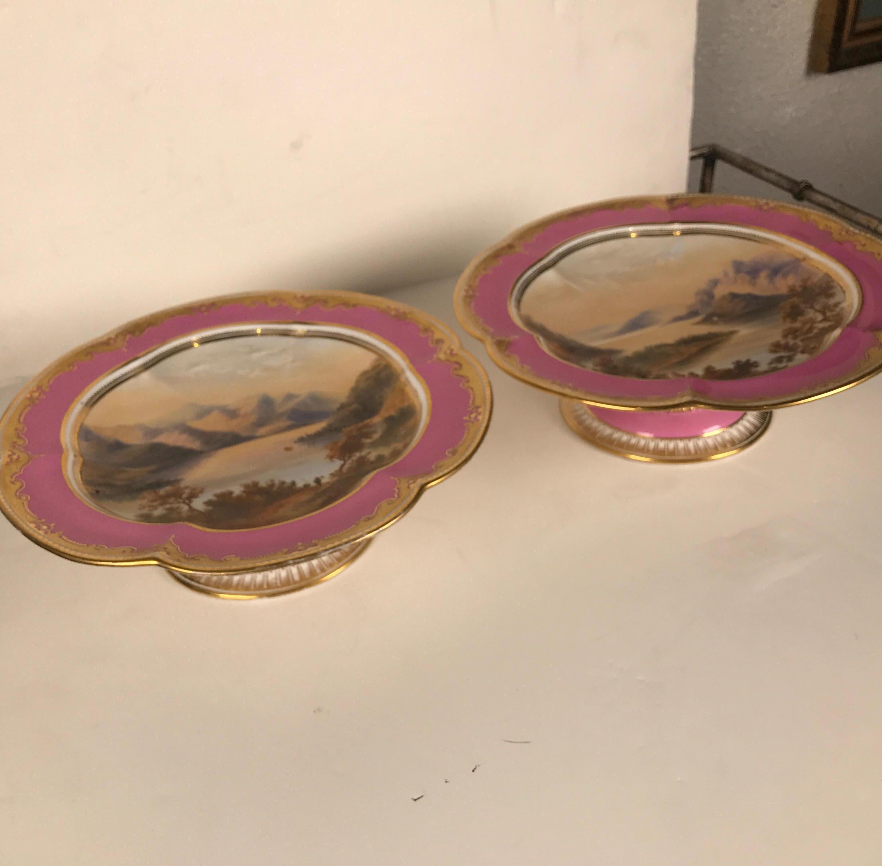 A pair of antique English hand painted and raised gilt porcelain compotes. Each one is painted a different Irish seaside landscape. The remarkable hand painted plates with raised gilt and lavender borders. The pedestal bases with gilt decoration.